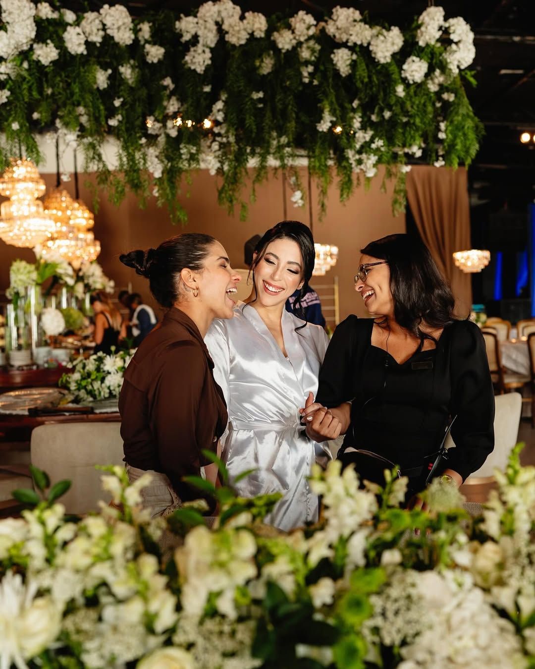 Three people laughing together in a decorated event space, with white florals in the foreground and hanging above.
