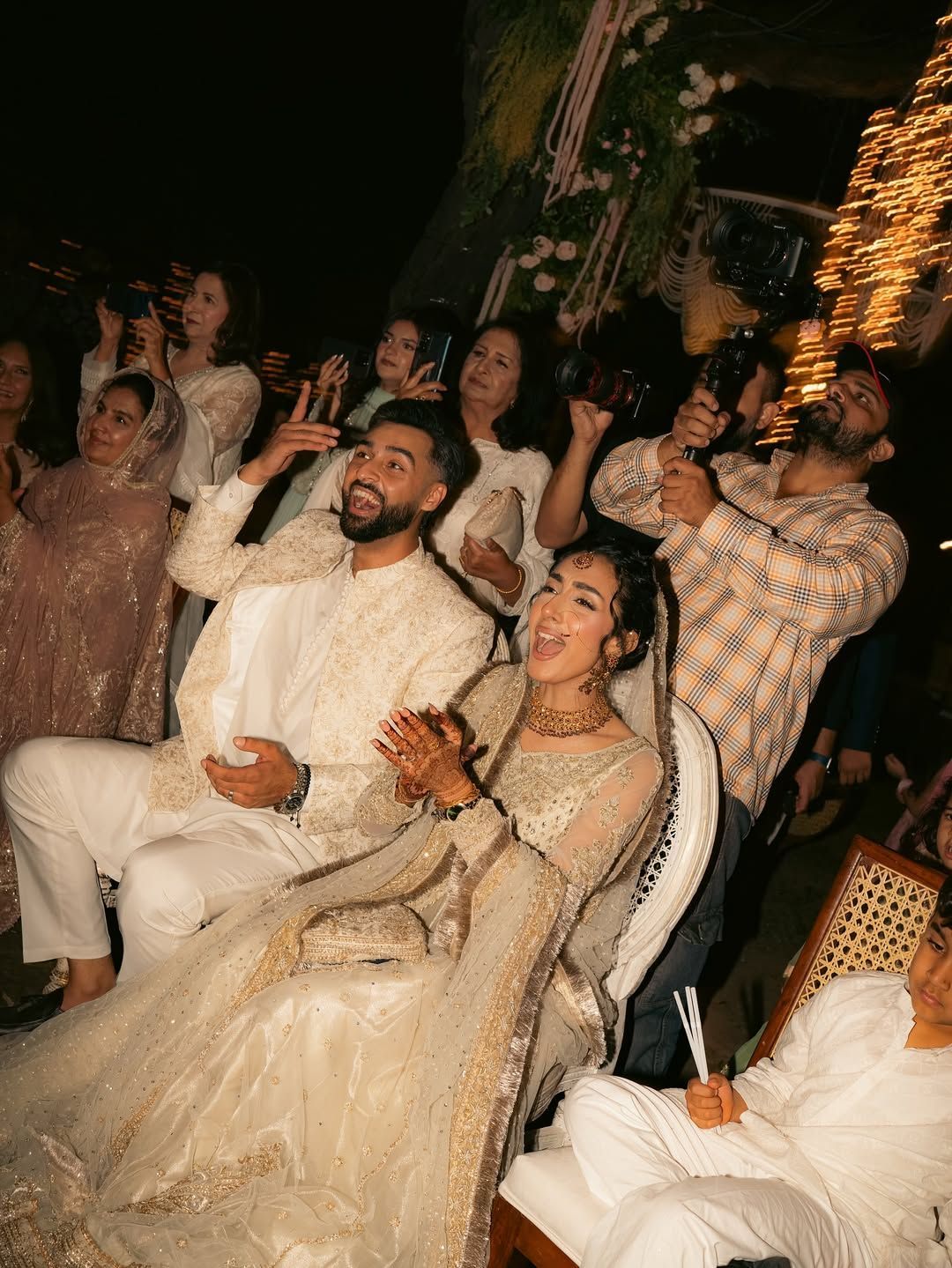 A couple in formal, embroidered cream attire sits and celebrates, surrounded by guests looking upward at an event.