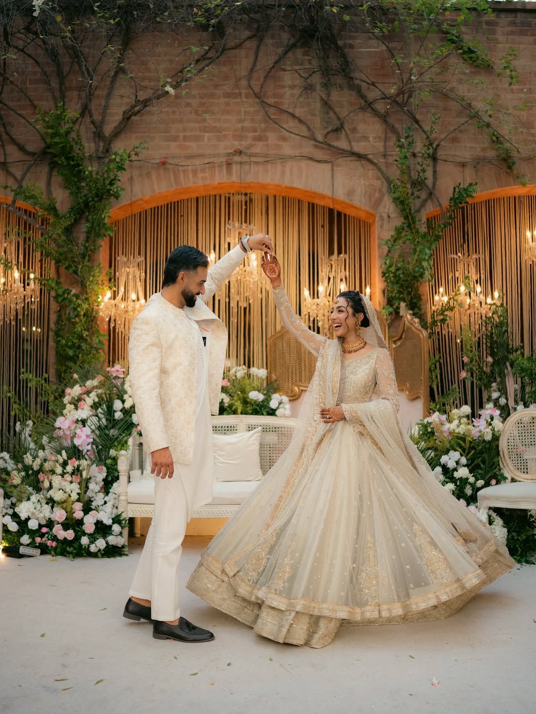 A couple in cream-colored formal wedding attire dance in a floral, candlelit outdoor venue.