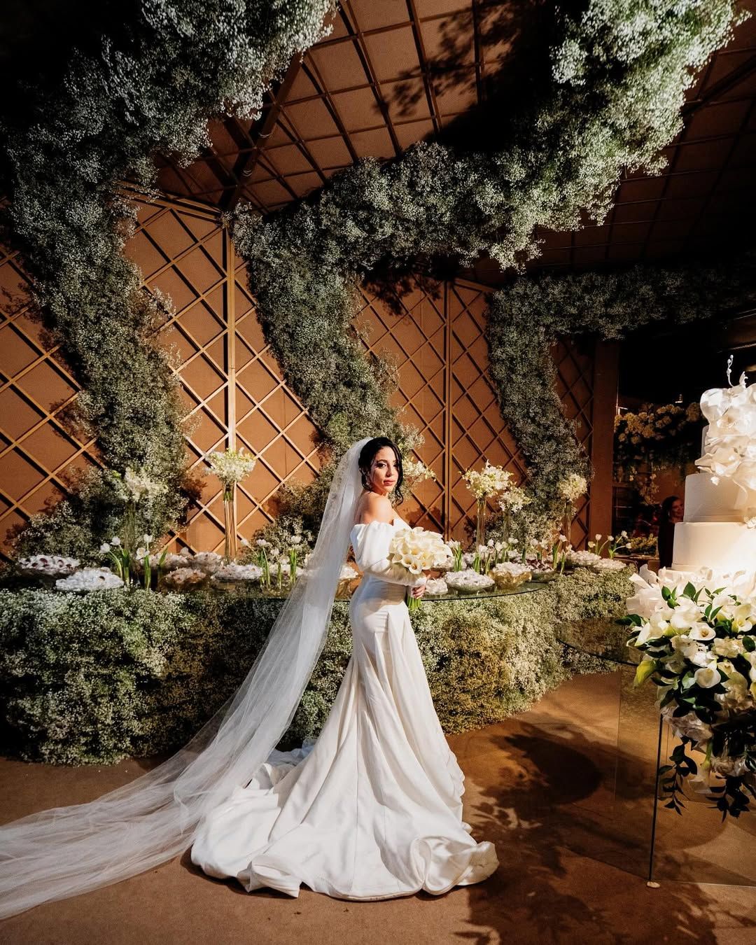 A bride in a white gown and veil stands centered in an elegant, flower-adorned venue with a decorative lattice wall.