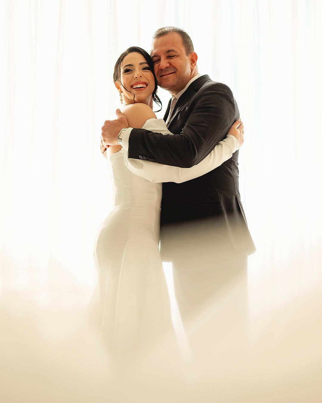 A couple embracing warmly, dressed in formal wedding attire, stands before a bright, softly lit window.