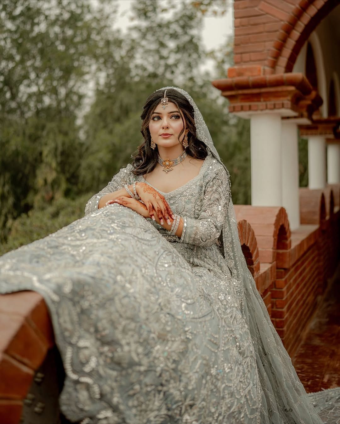 A person in a heavily embellished silver gown and bridal veil poses elegantly on a brick balcony outdoors.