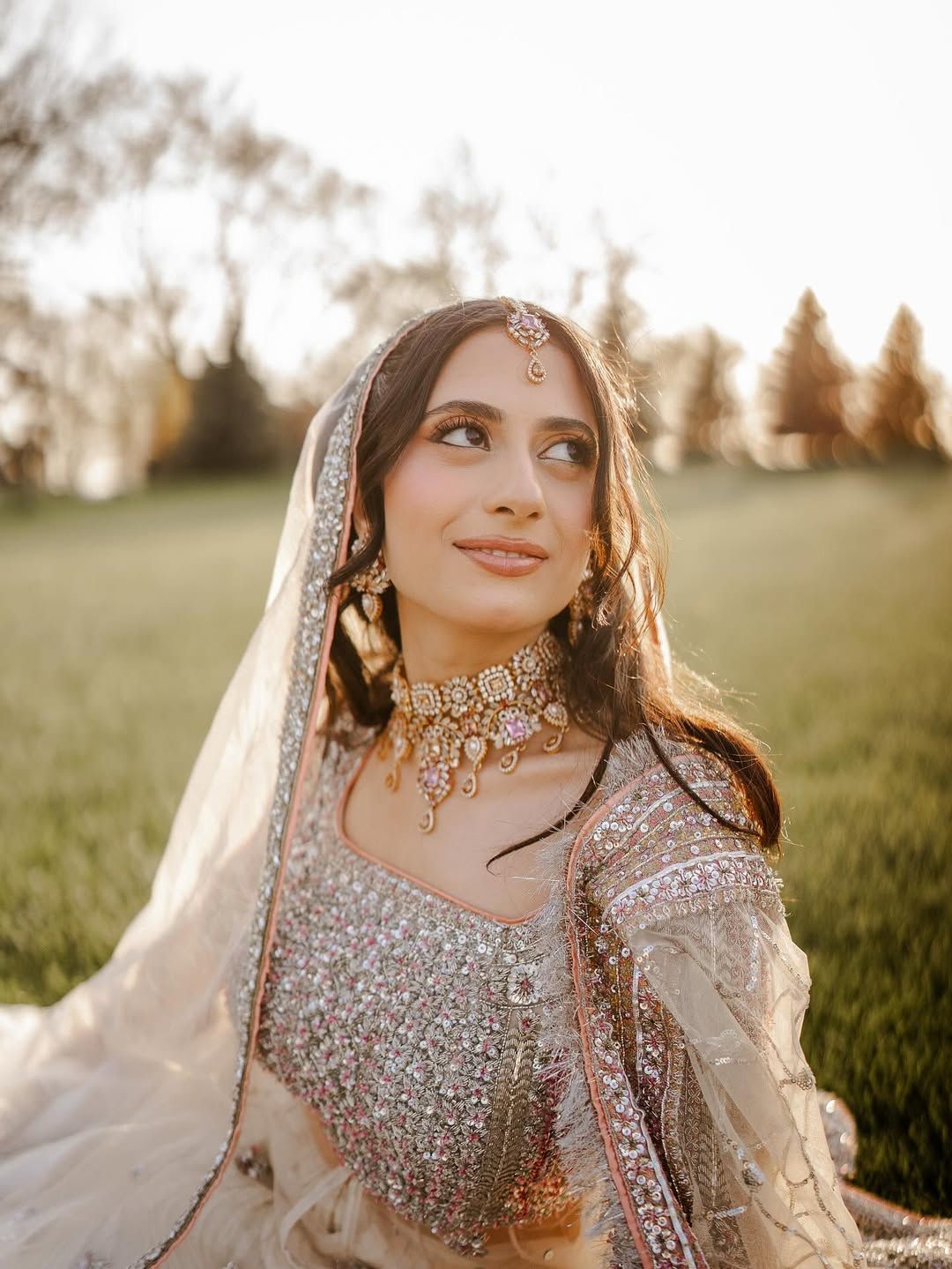 A person wearing an ornate, sparkling bridal lehenga and traditional jewelry poses in a grassy field at sunset.