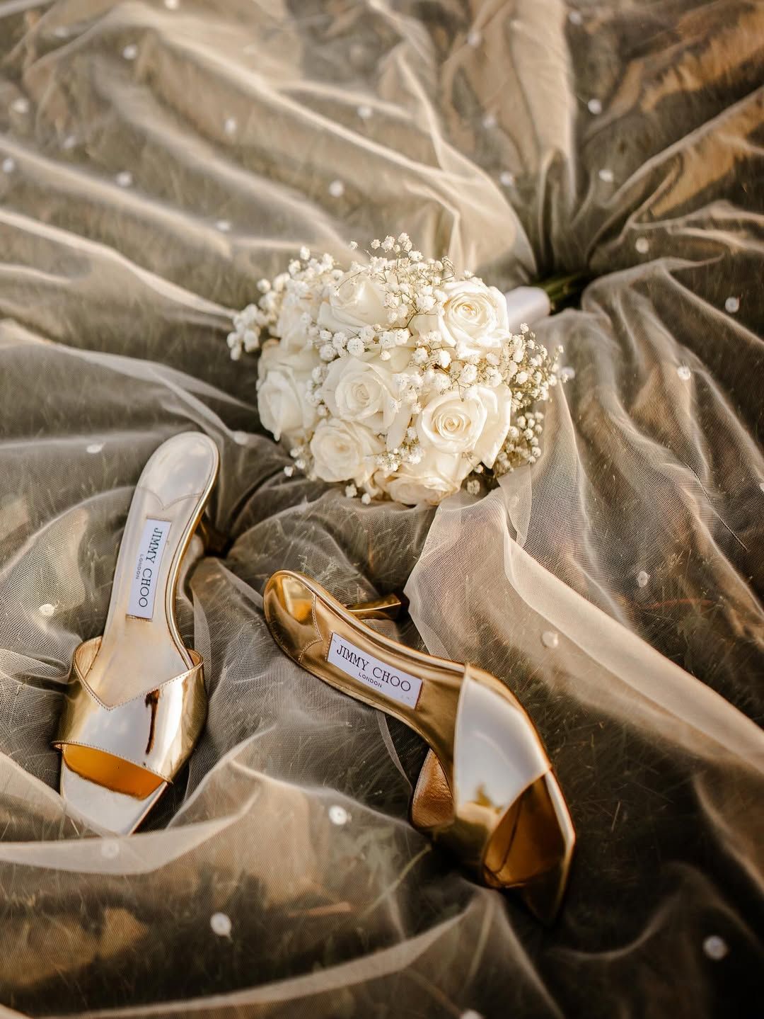 A pair of metallic gold heels and a white rose bouquet resting on a shimmering, polka-dot tulle fabric.
