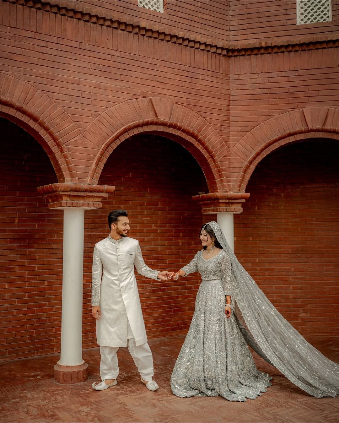 A couple in traditional white and silver wedding attire holding hands, posing under a red brick archway.