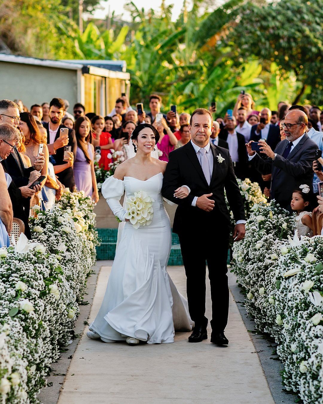 A couple walks down an aisle lined with white flowers at an outdoor wedding, surrounded by guests in a garden setting.