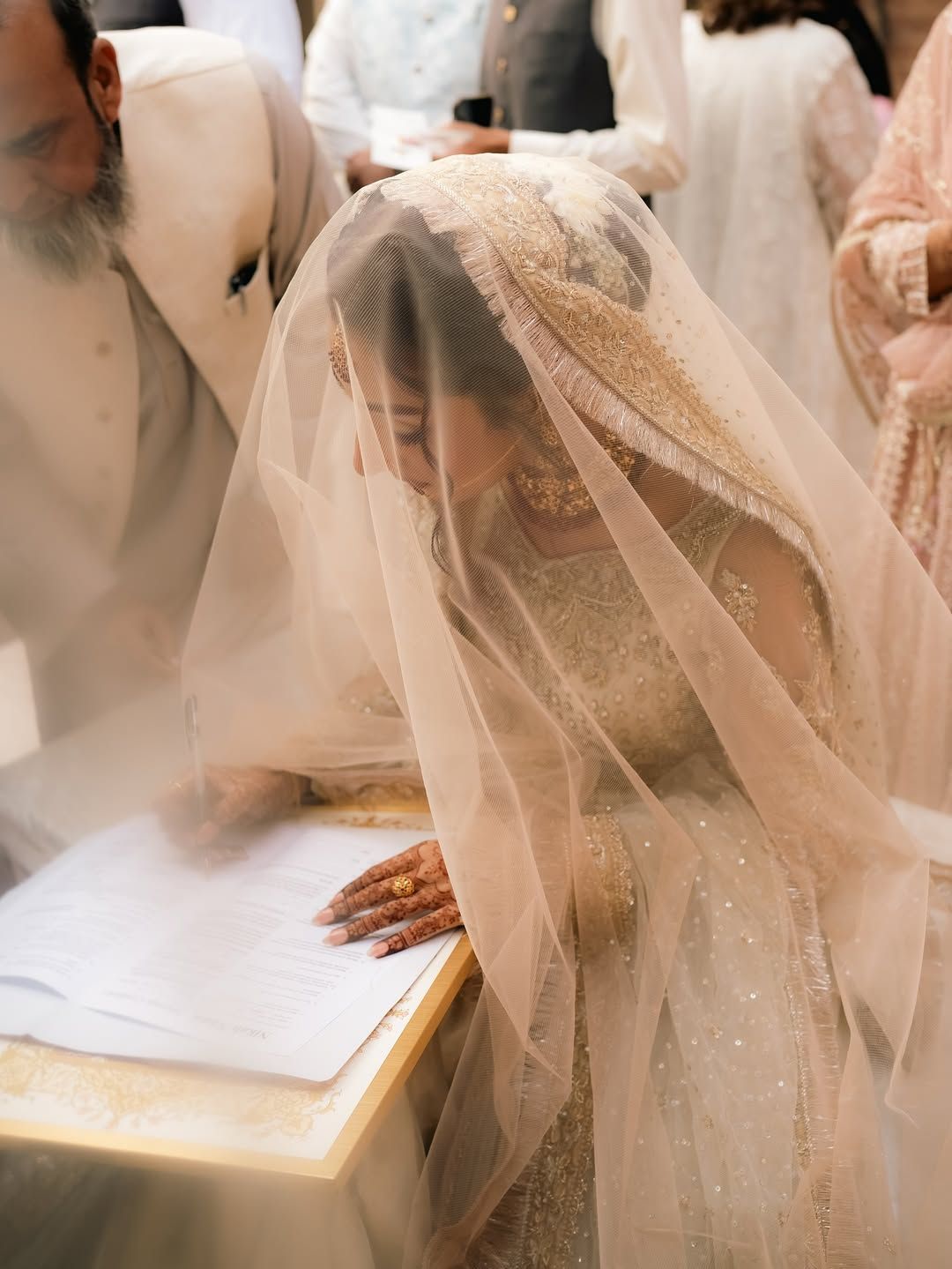 A person in an ornate, neutral-toned bridal outfit and veil signs a document during a ceremony.