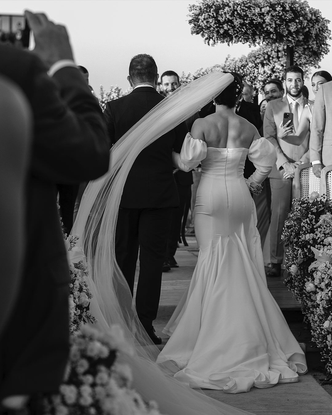 A bride and groom walk down an aisle at an outdoor ceremony, their backs to the camera, with guests watching on the sides.