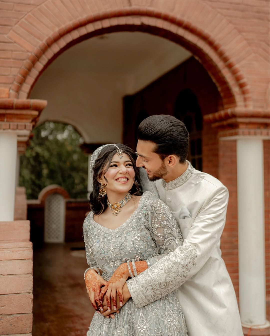 A couple in formal, intricately embroidered light-colored attire embracing under a brick archway.