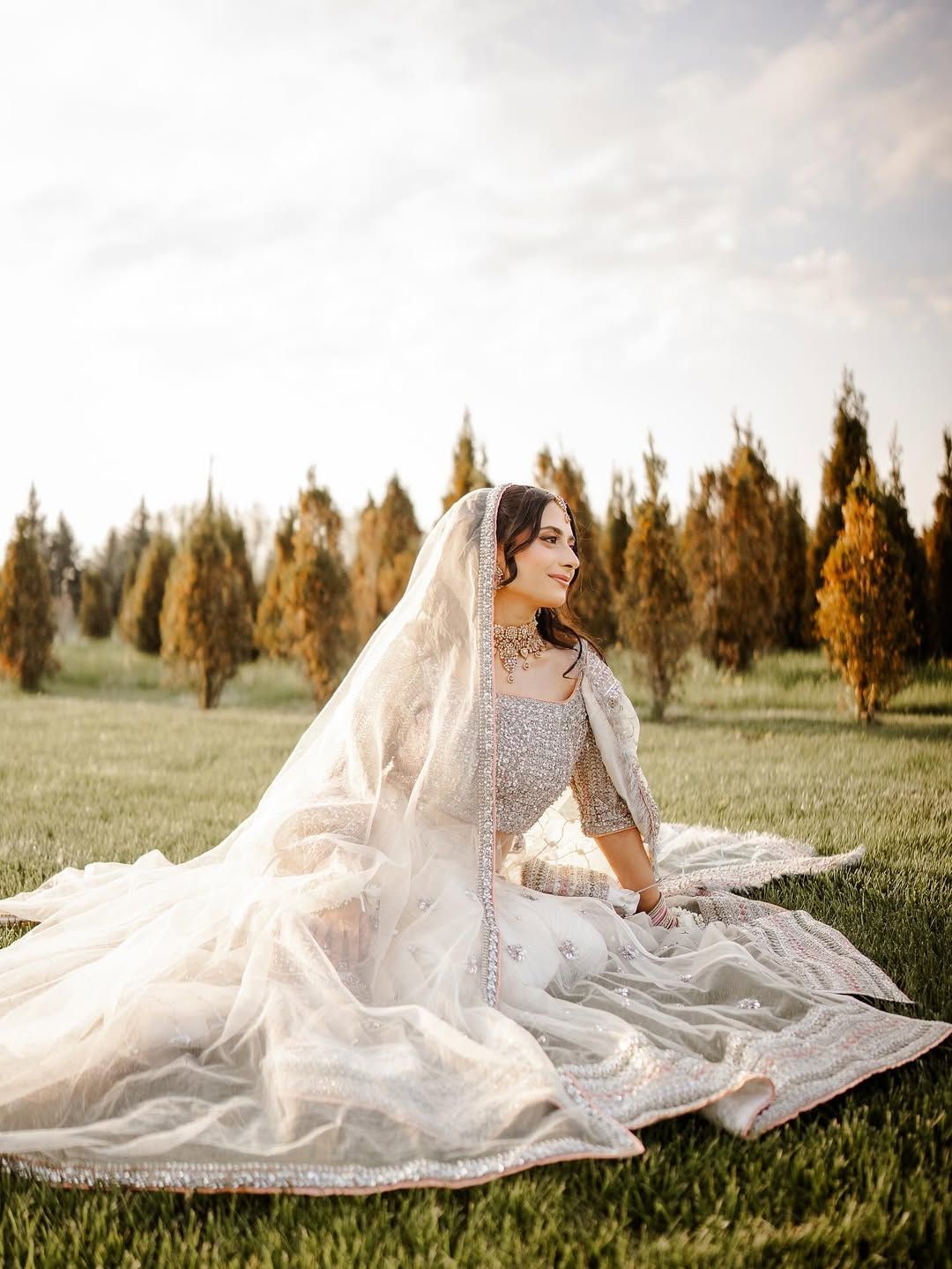 A person in a heavily embroidered white bridal outfit and veil sits gracefully in a grassy field among evergreen trees.