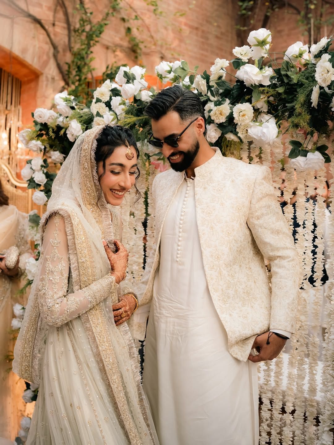 A couple in formal, ivory wedding attire stands together smiling under a floral archway.