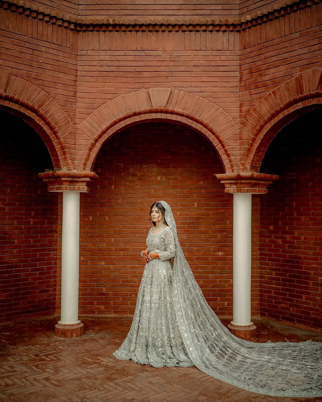 A bride in a long, silver-embroidered gown and veil stands between white columns in a rustic, arched red-brick corridor.
