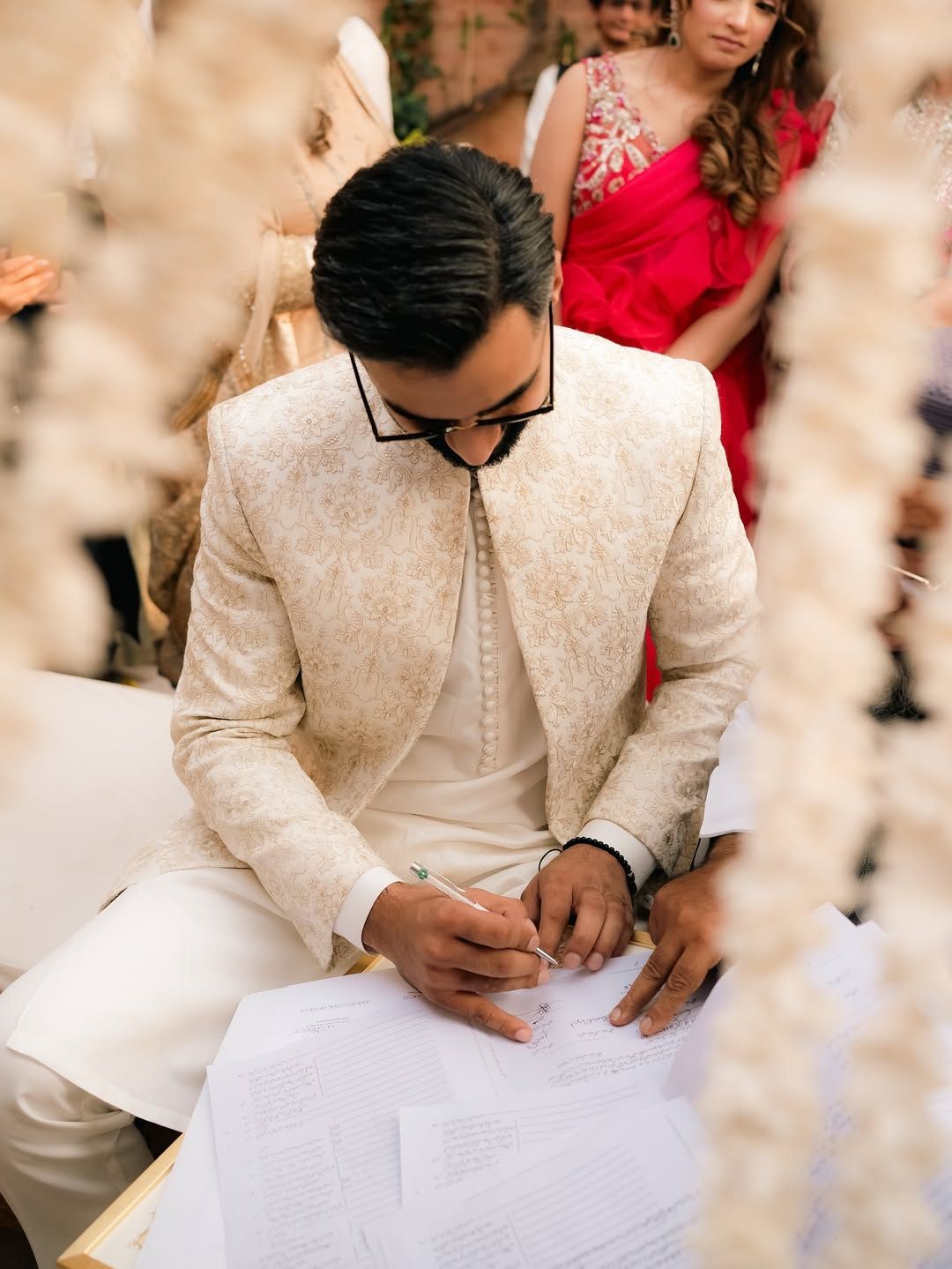 A person wearing a cream-colored traditional sherwani signs a document at a wedding ceremony, surrounded by guests.