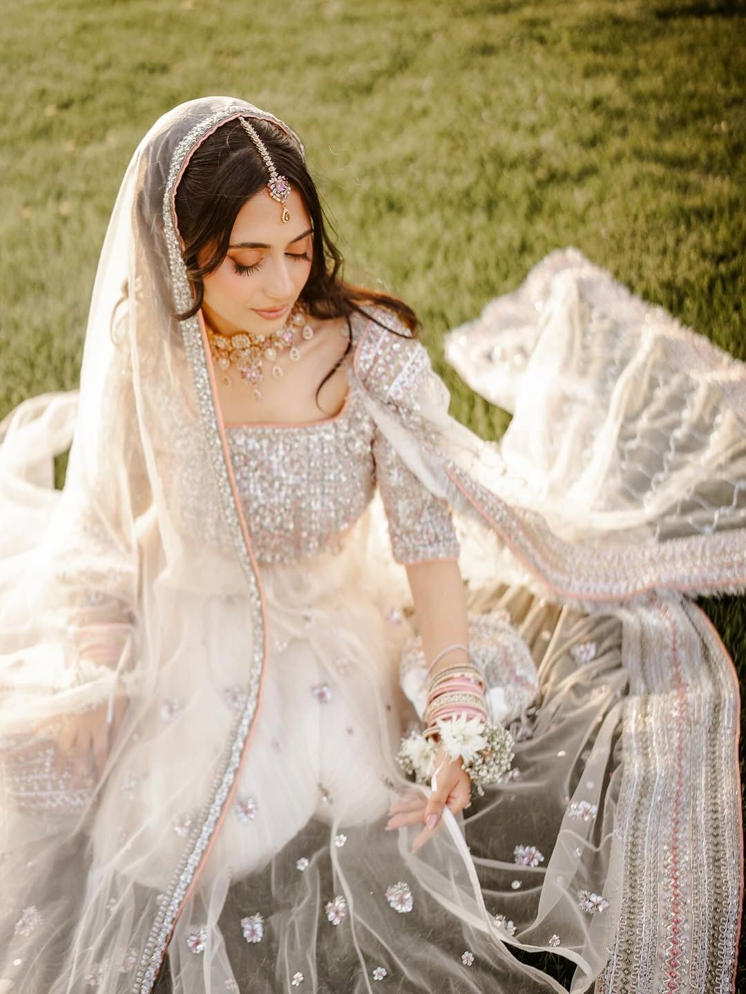 A person in an ornate, silver-embellished traditional white bridal outfit and veil, sitting gracefully on green grass.
