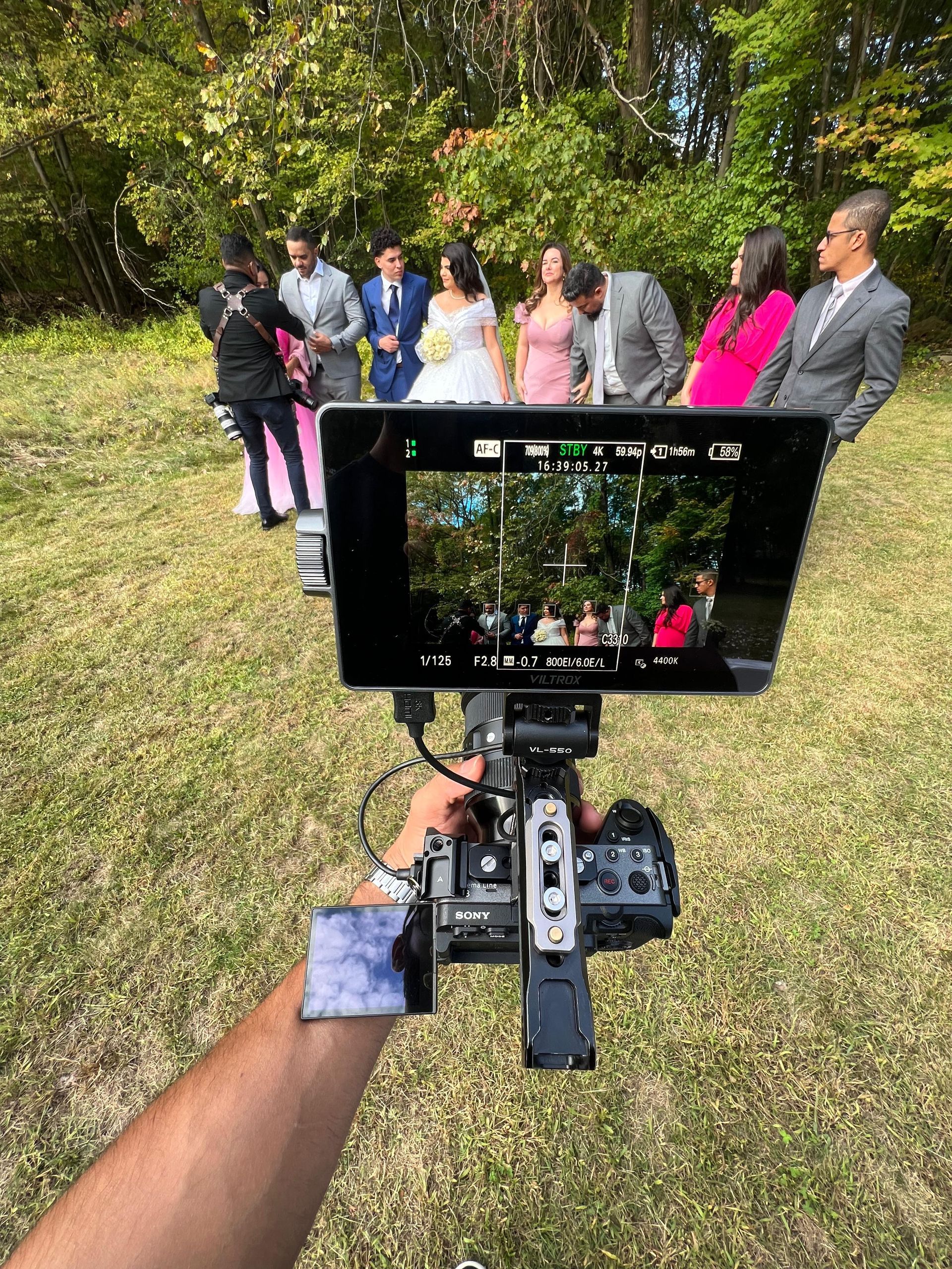 A first-person view of a camera filming a wedding party standing together outdoors on a grassy lawn.