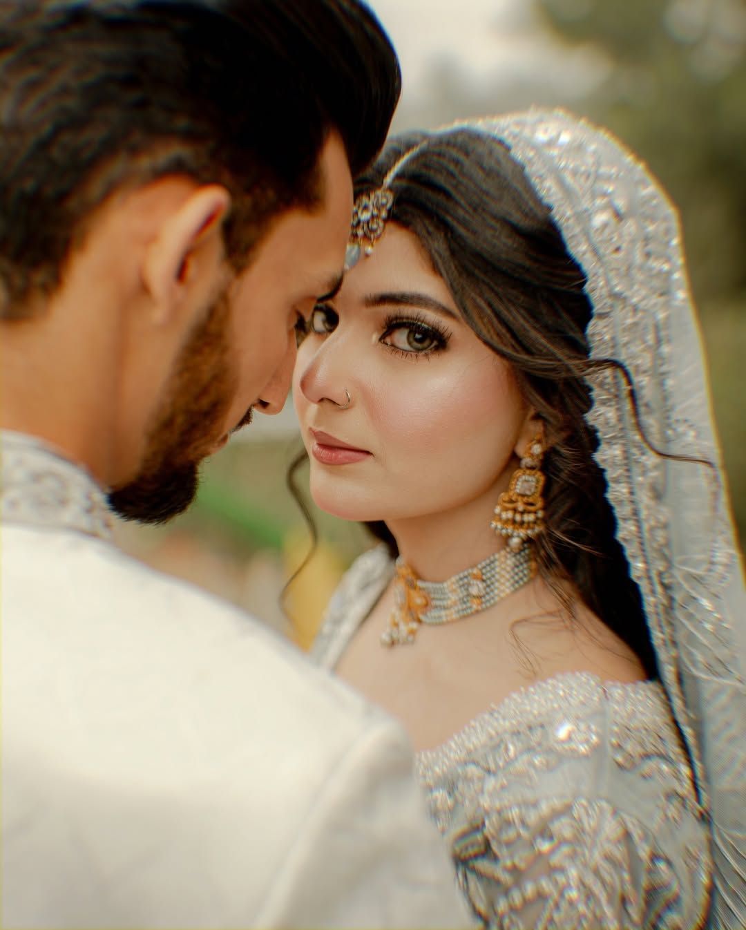 A couple in formal, intricately embroidered pale wedding attire stand closely, looking at each other in a soft, bright set.