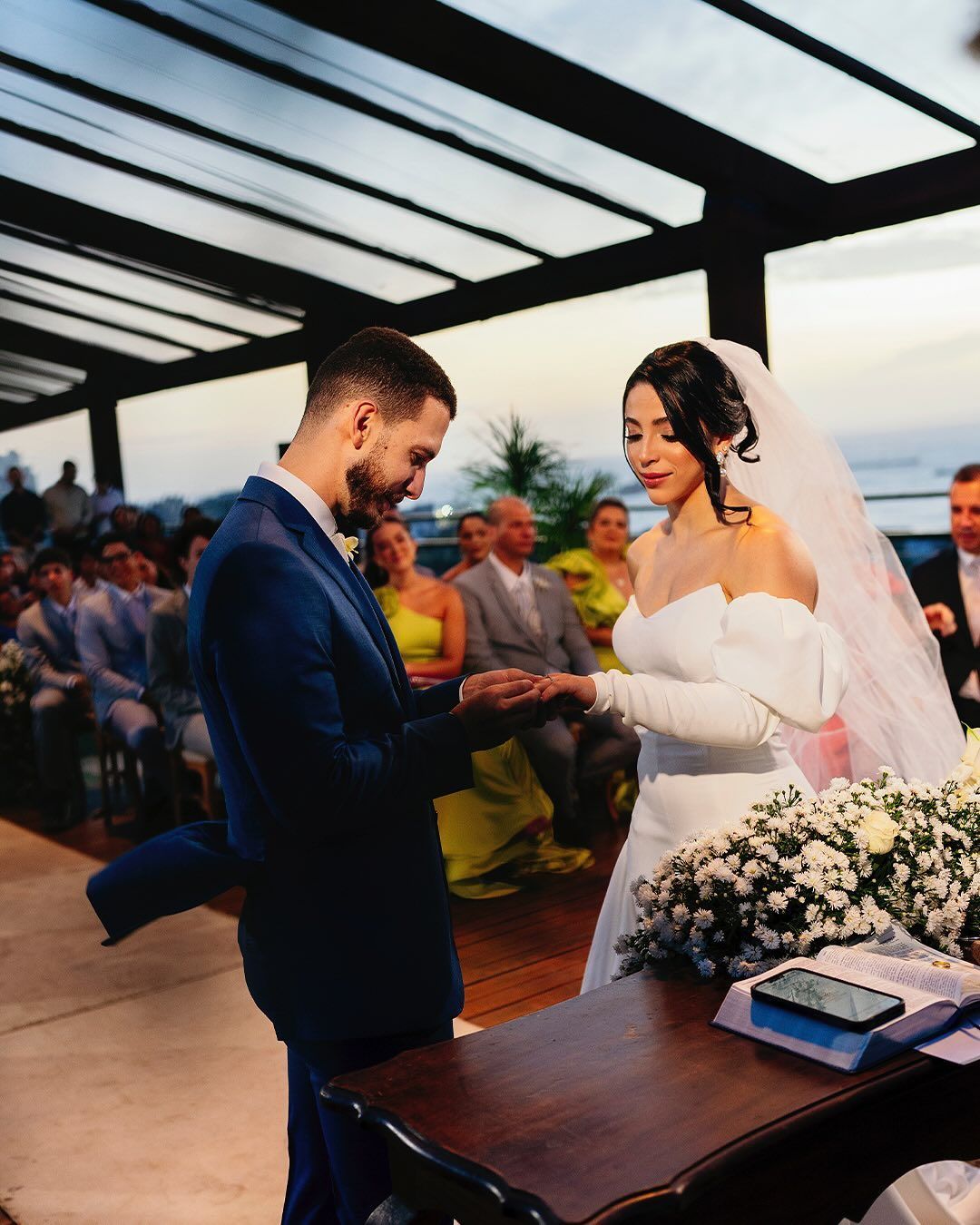 A bride and groom exchange rings during a wedding ceremony on an outdoor terrace at dusk.
