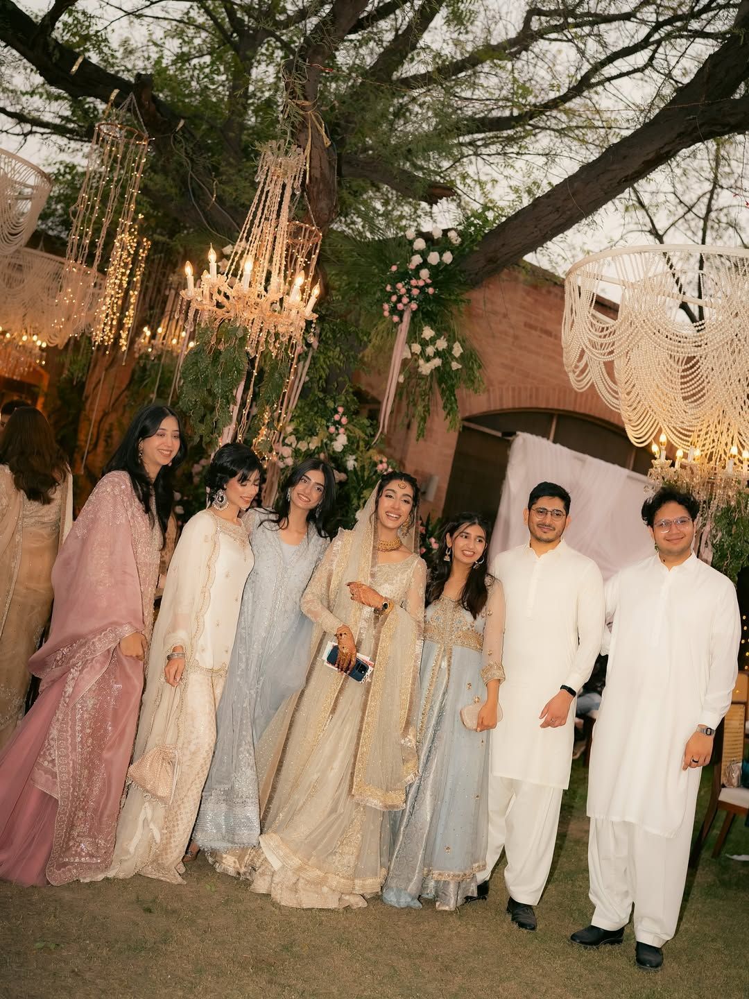 A group of people in formal South Asian attire pose for a photo at an outdoor evening event with hanging chandeliers.