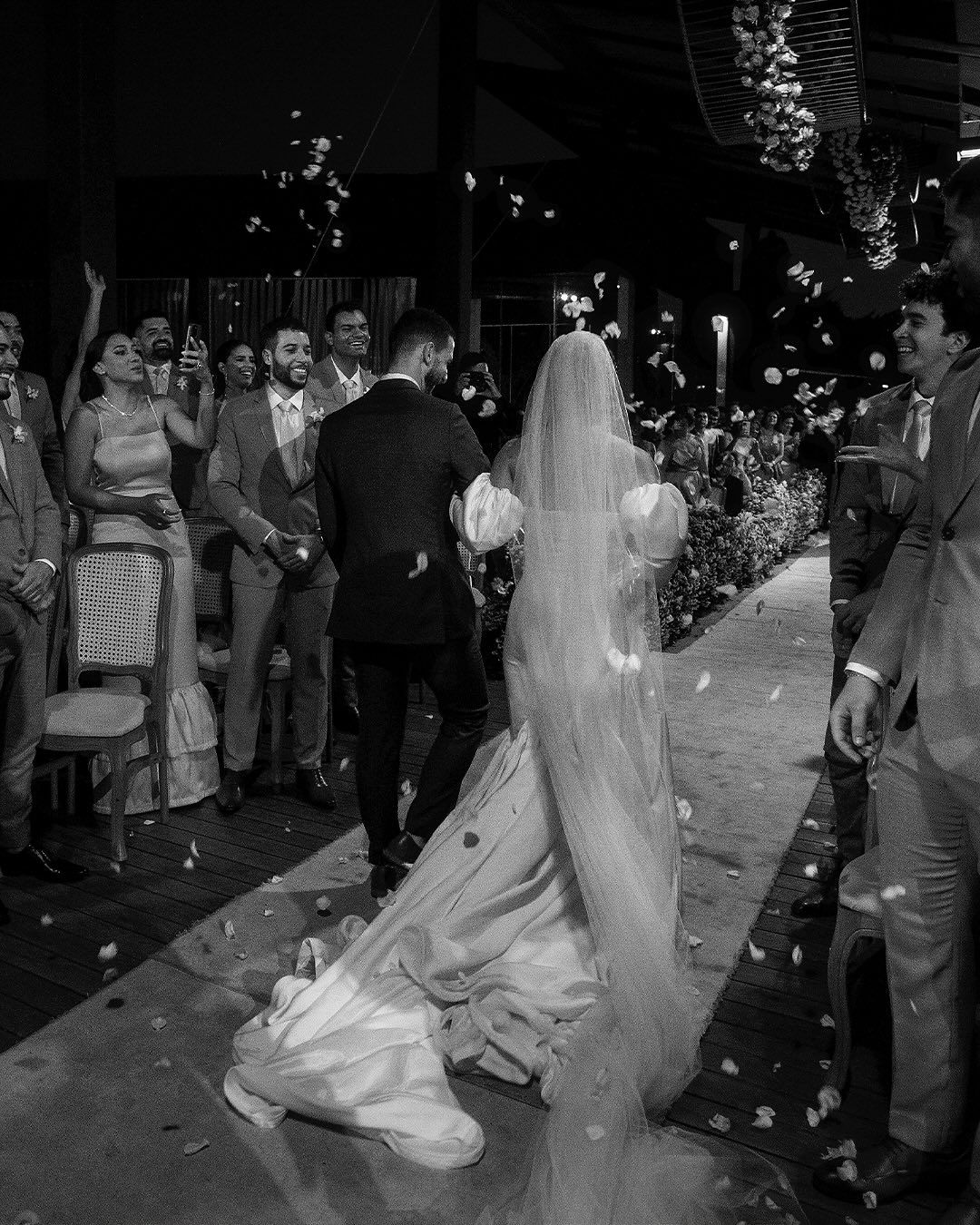 A bride and groom walk down an aisle as guests throw flower petals in a black and white wedding scene.