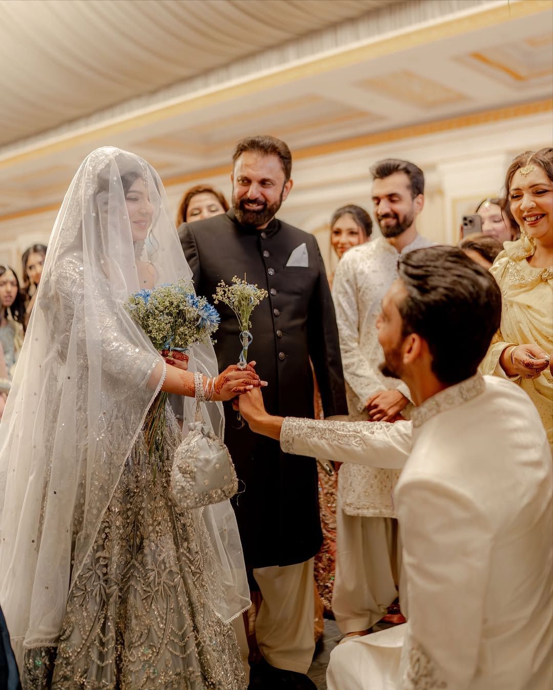 A person in traditional wedding attire kneels, taking the hand of a bride, surrounded by guests in a formal setting.