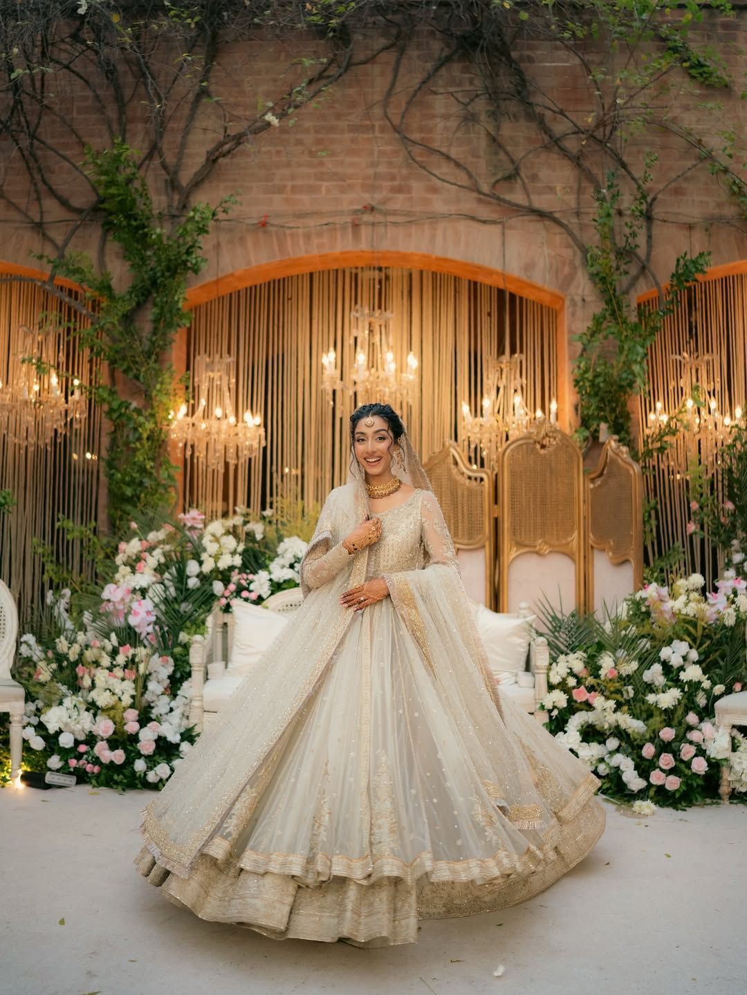 A person in a cream-colored, heavily embroidered wedding dress standing in front of floral arrangements and chandeliers.