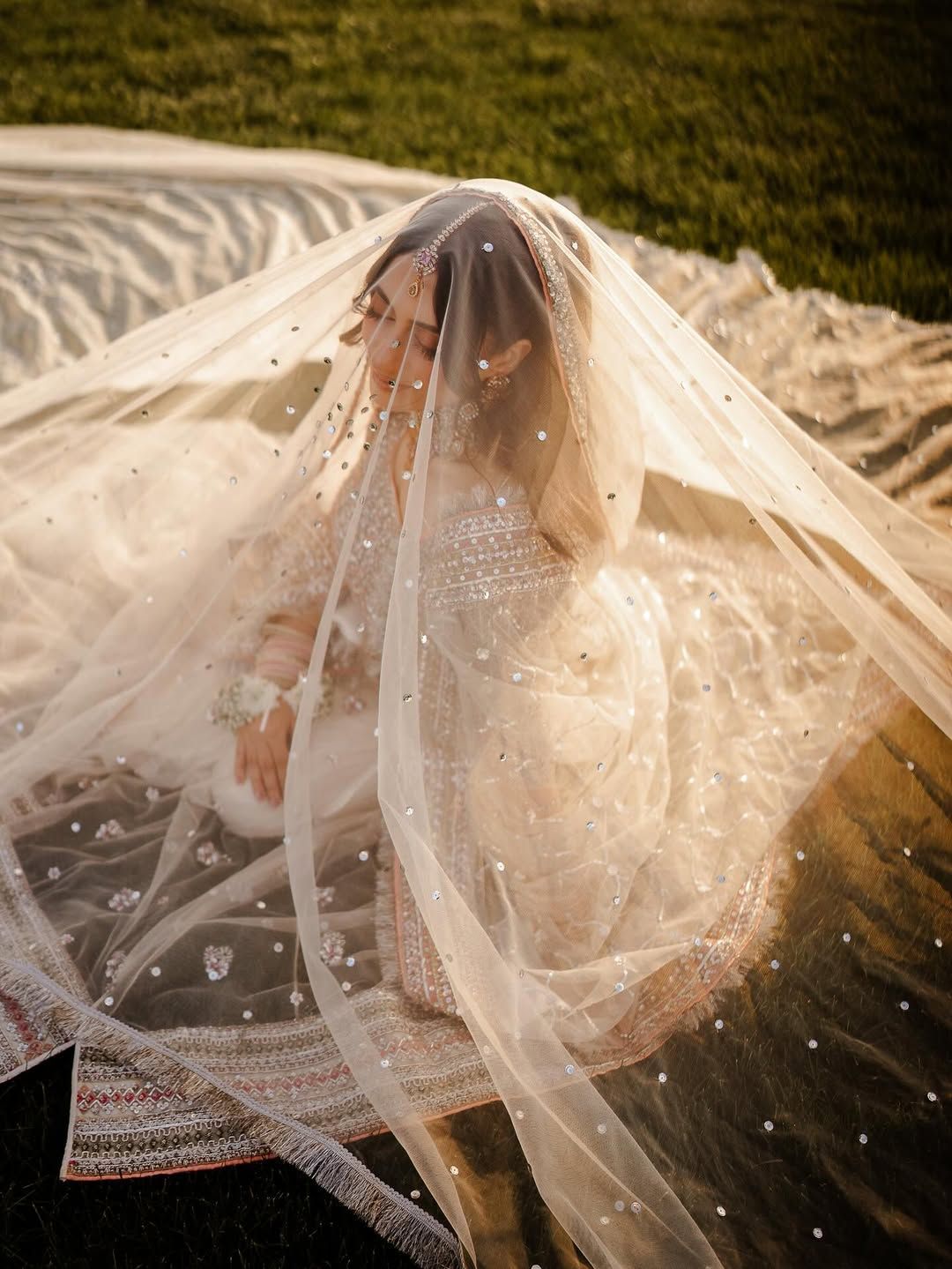 A person in a beaded wedding dress sits on grass, draped in a sheer, floor-length veil adorned with scattered sparkles.