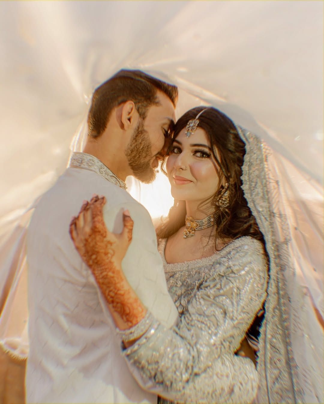A couple in ornate, light-colored wedding attire embrace under a semi-transparent veil, bathed in soft, warm golden light.