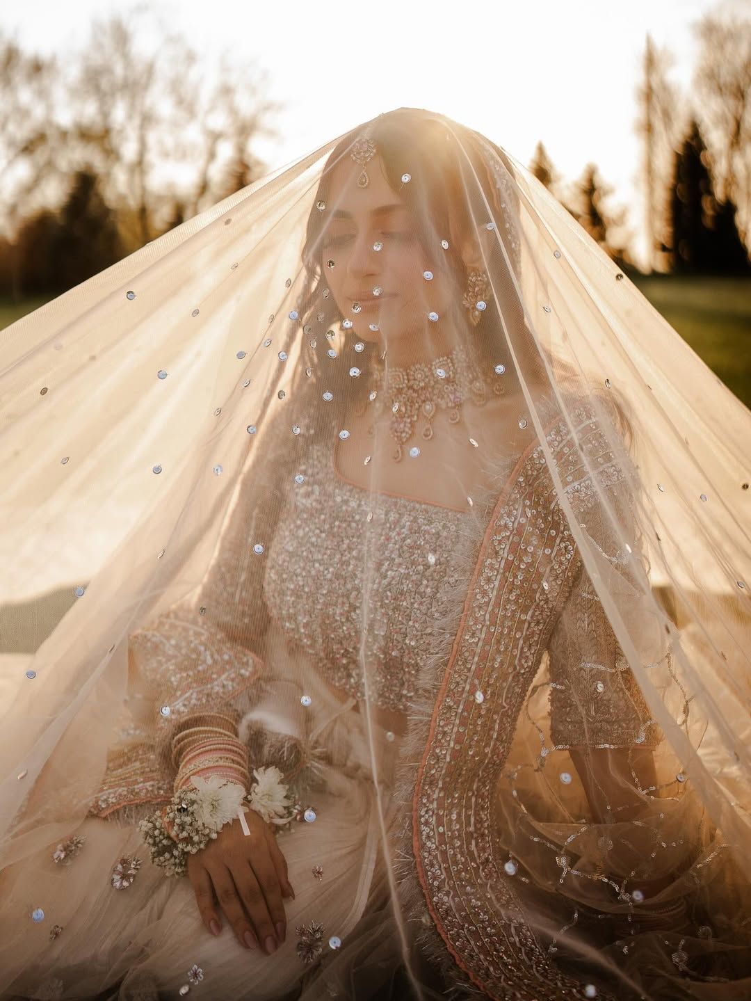 A bride wearing a heavily beaded gown and veil stands in soft, golden sunlight.