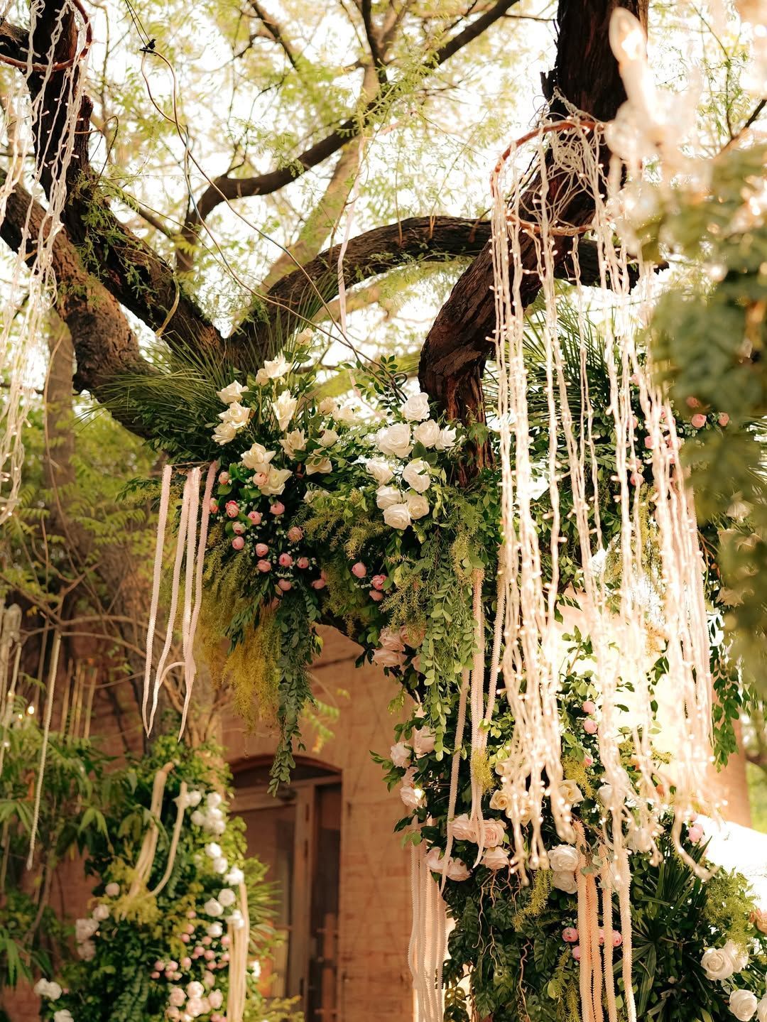 A tree decorated with hanging white ribbons, lush green foliage, and white flower arrangements for an outdoor event.