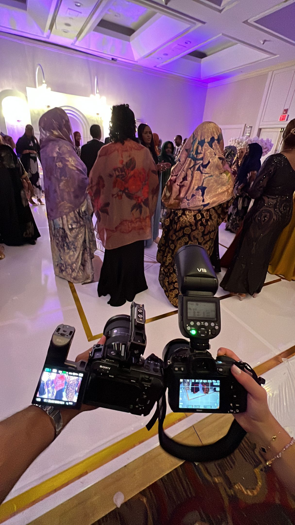 A photographer’s perspective holding two cameras at a banquet hall event with guests in traditional attire in the back.