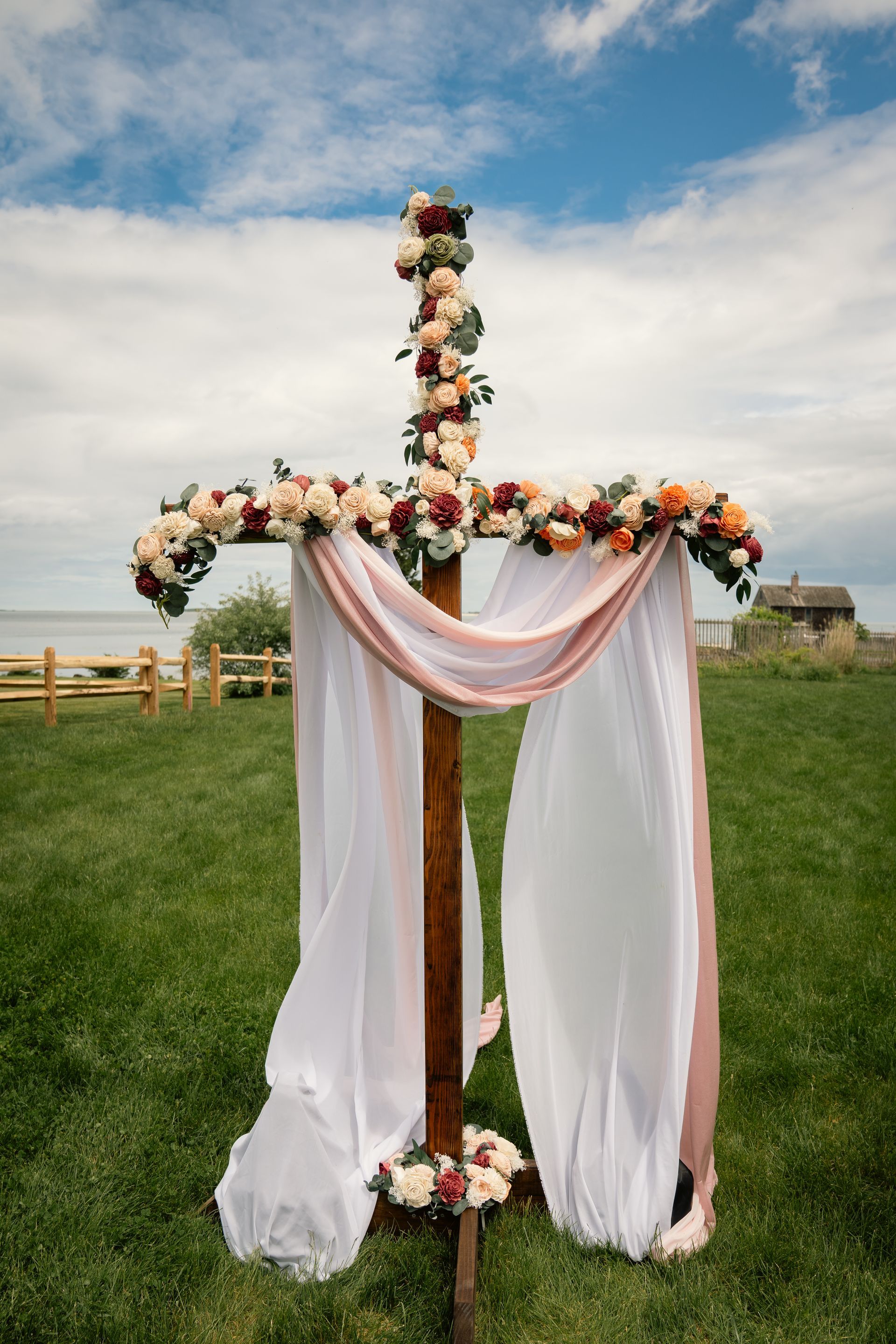 A wooden cross wedding arch decorated with cream, peach, and burgundy flowers, draped with white fabric in a grassy field.