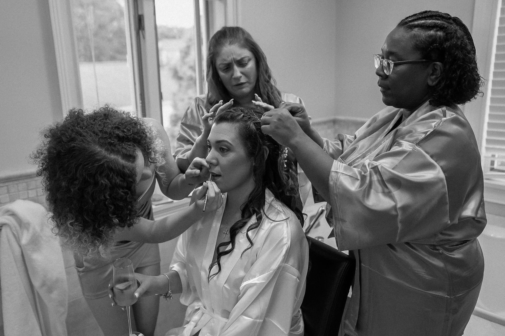 Three people helping a bride get ready for a wedding in a room, styling her hair and applying makeup.