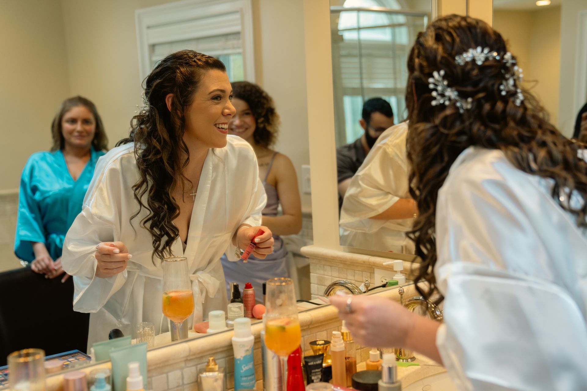 A person in a white robe smiles at their reflection in a bathroom mirror while other people stand in the background.