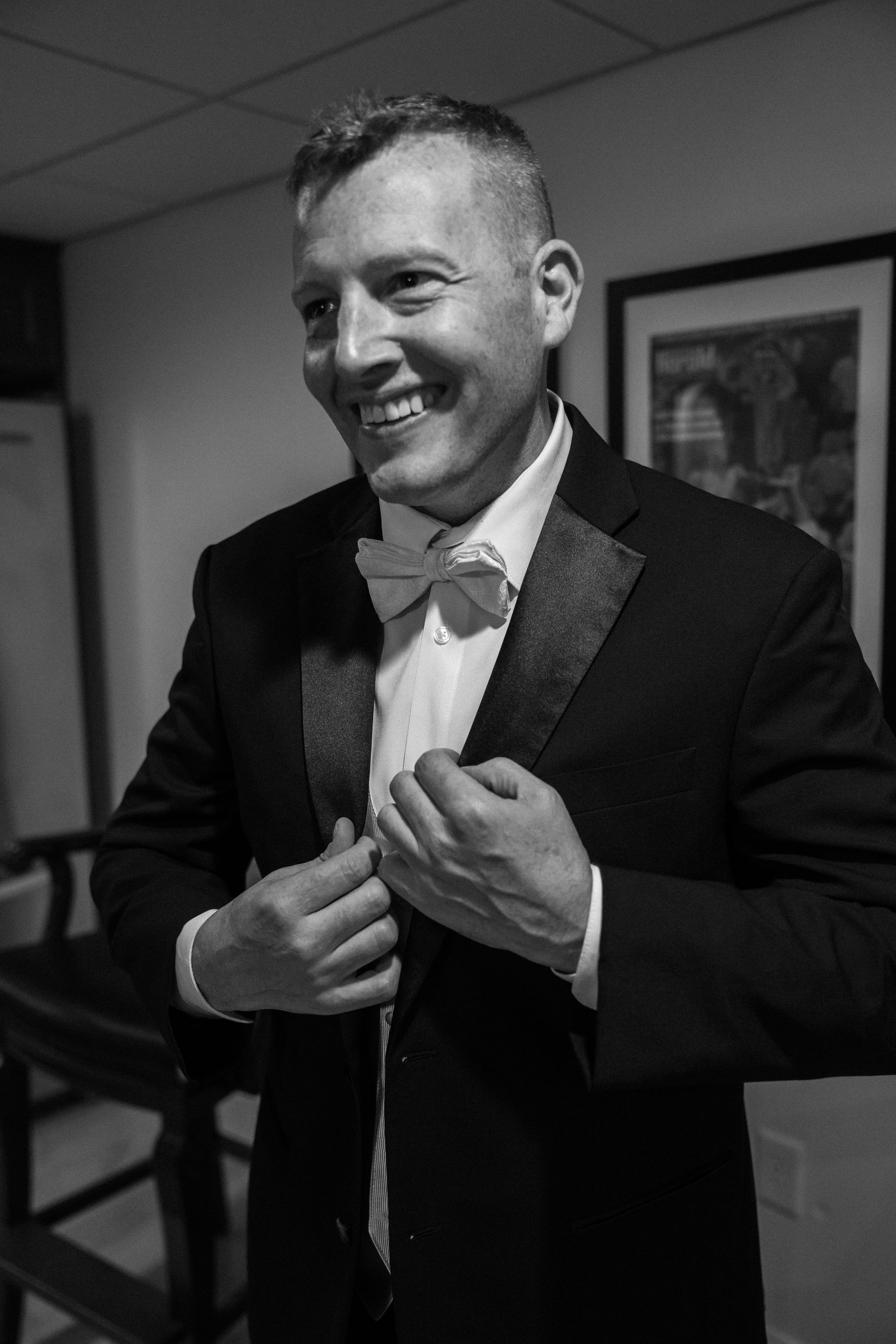 A person in a black tuxedo with a bowtie smiles while adjusting their lapel in an indoor setting.