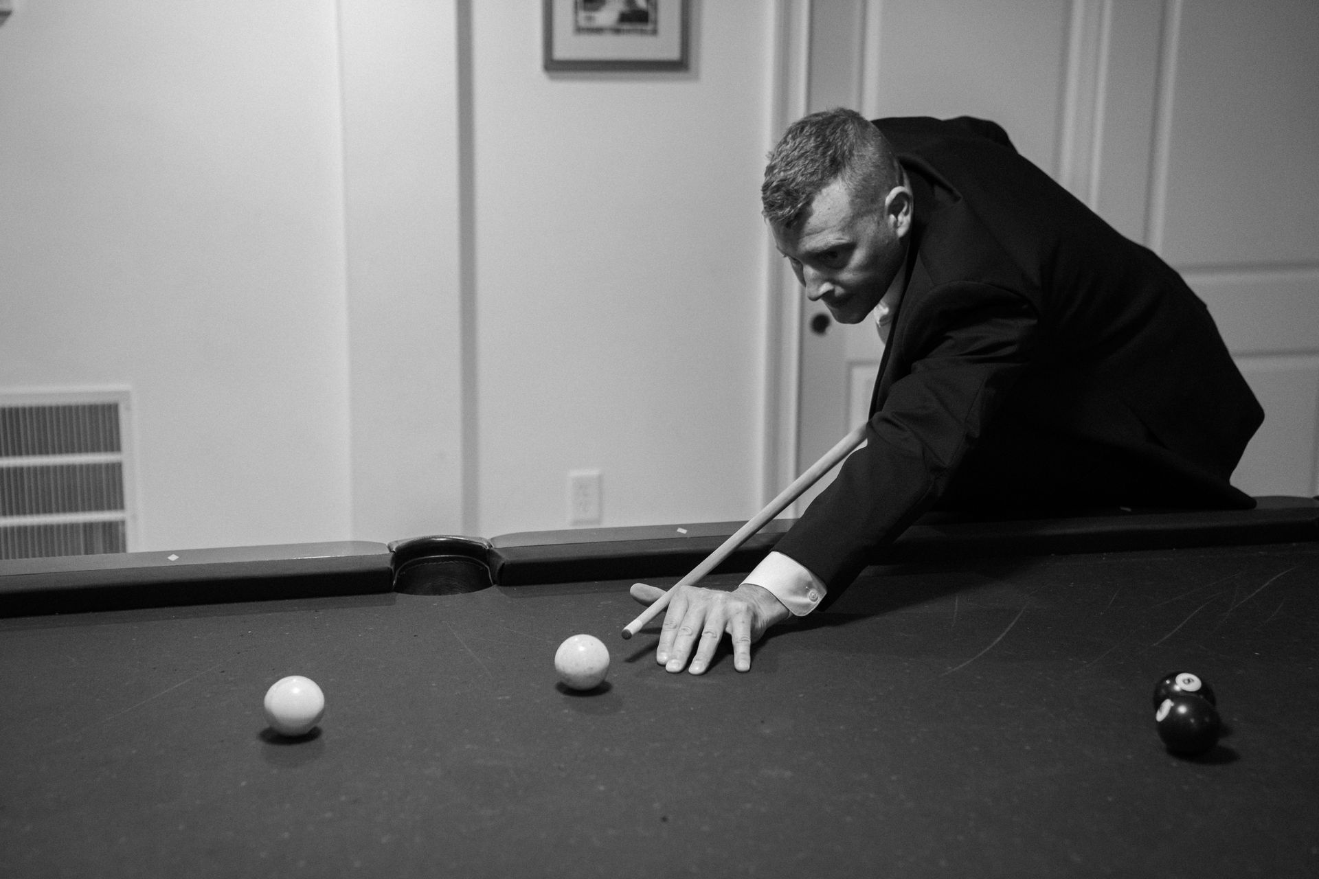 A person in a suit leans over a pool table, poised to strike a cue ball with a pool cue.