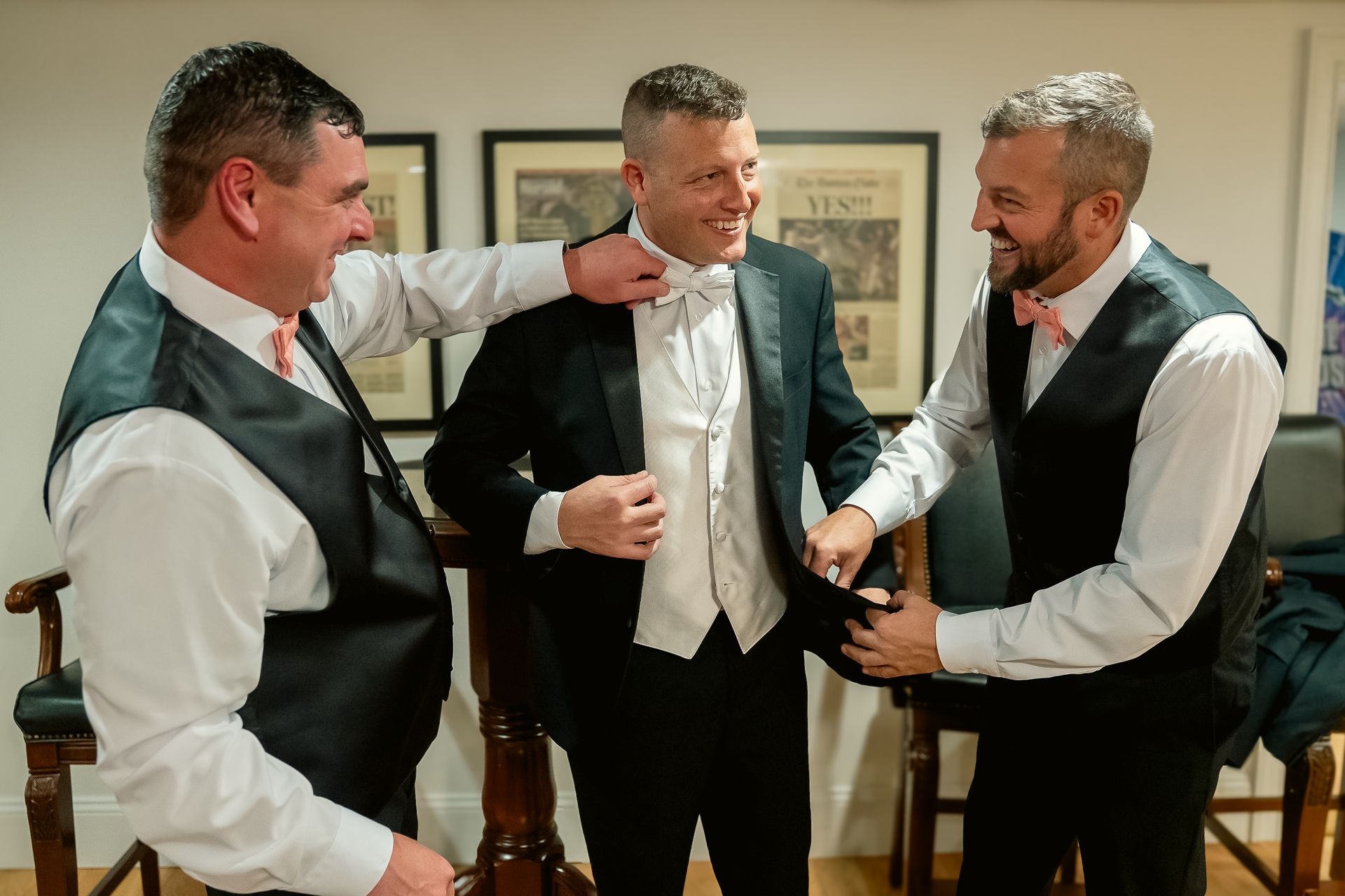 Three people in formal wedding attire laughing while helping one another adjust their bow ties and jackets.