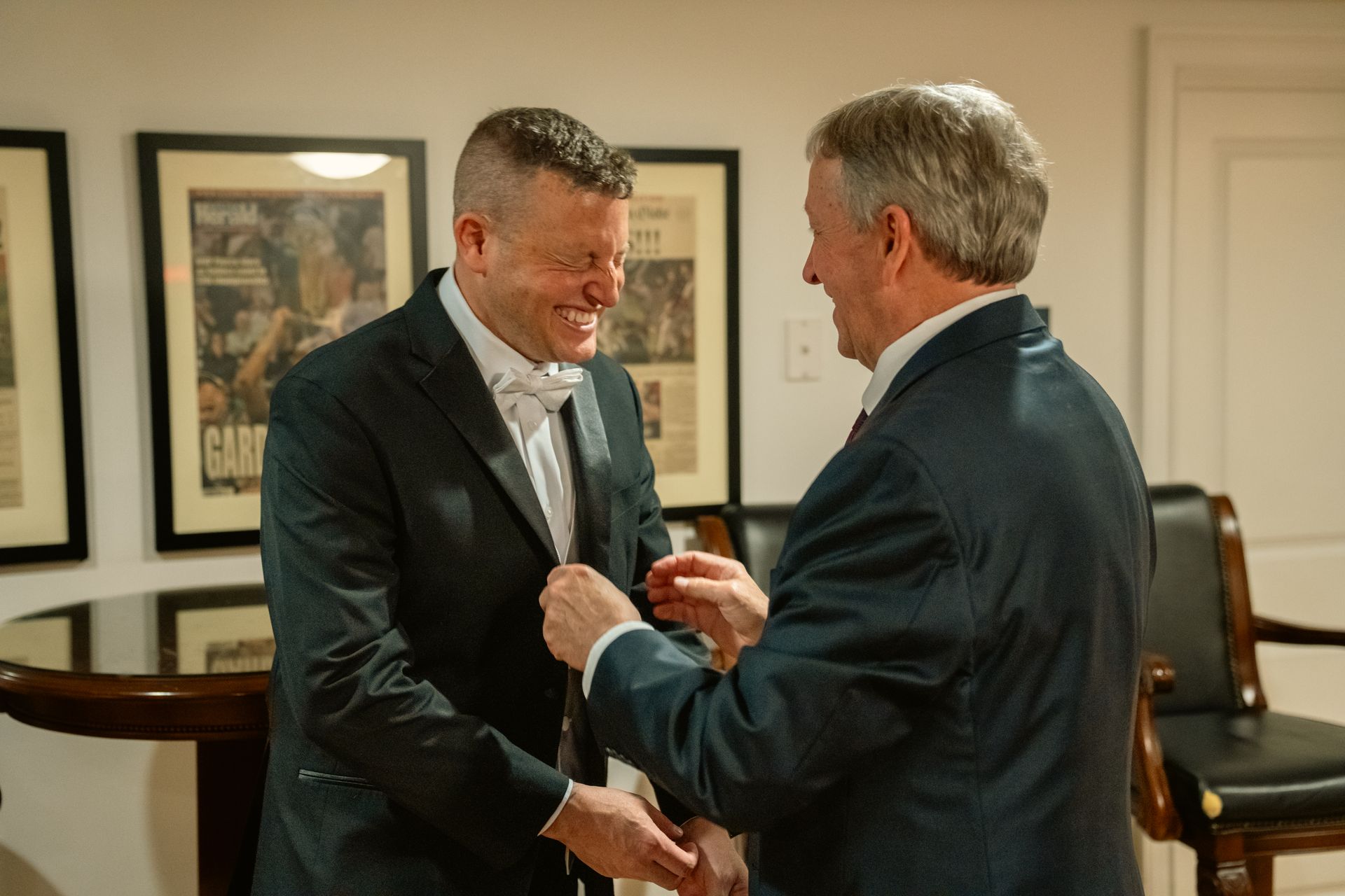 Two men in suits stand in a room, with one man adjusting the lapel of the other's jacket while both smile.