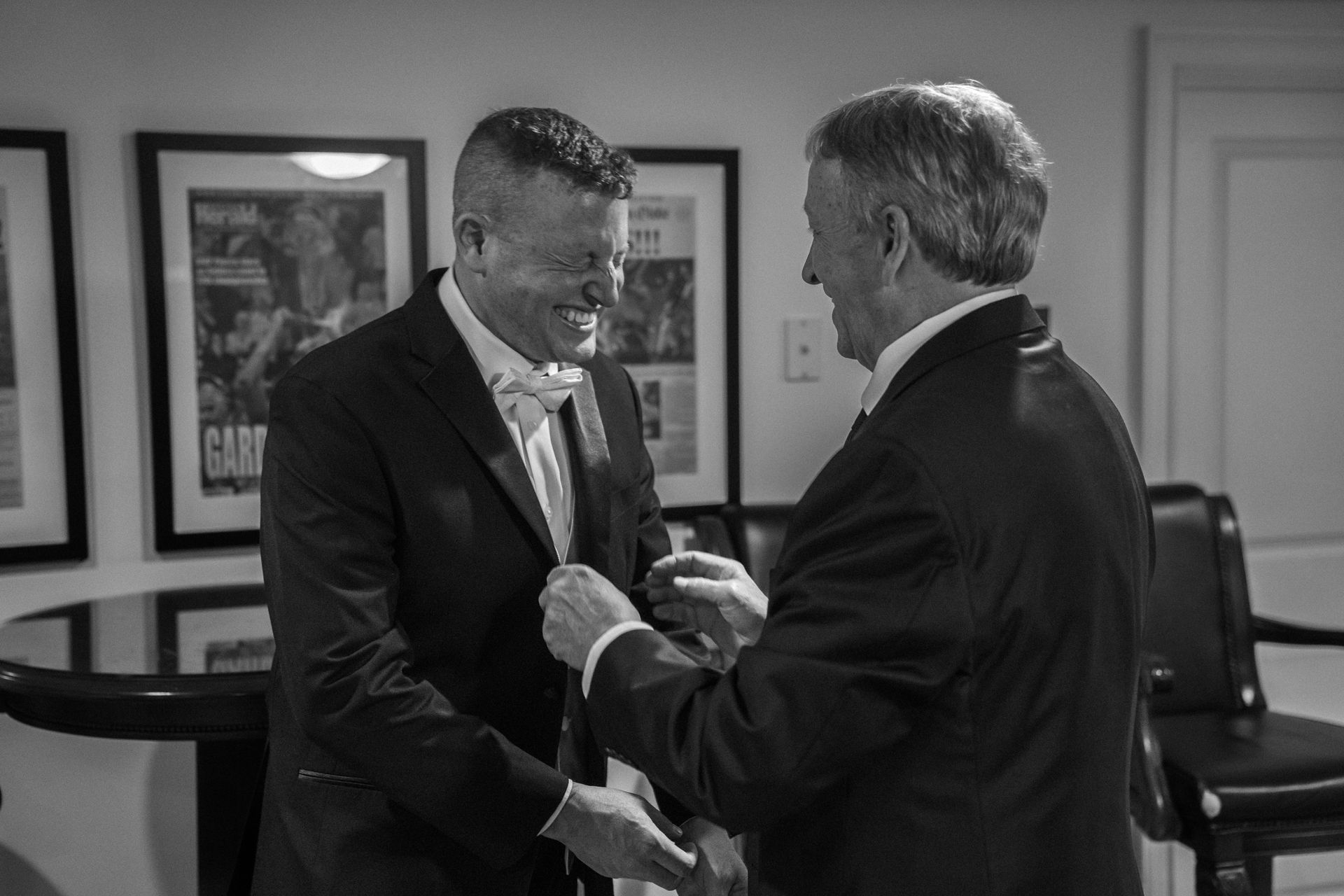 An older man adjusts the tie of a smiling man in a suit inside a room with framed pictures on the wall.