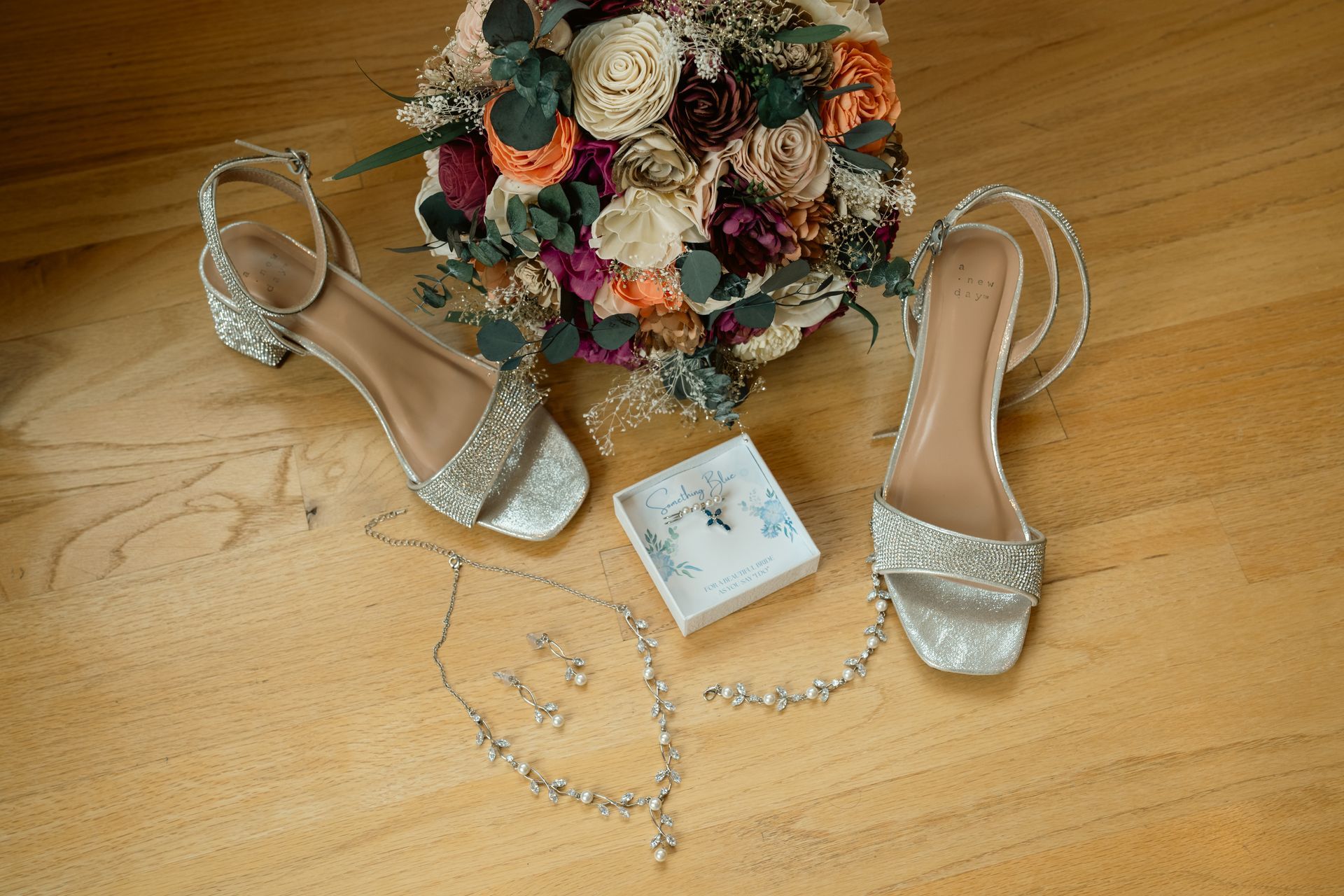 Wedding bouquet, silver embellished heels, and jewelry set arranged on a wooden floor.