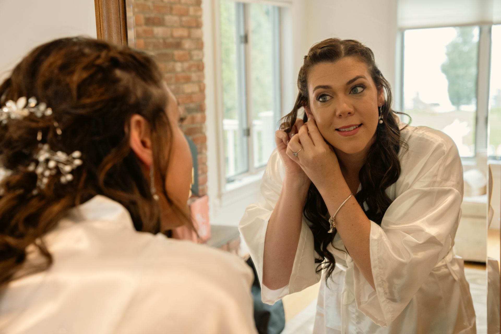 A person wearing a white robe looks into a mirror to put on an earring while preparing for a wedding.