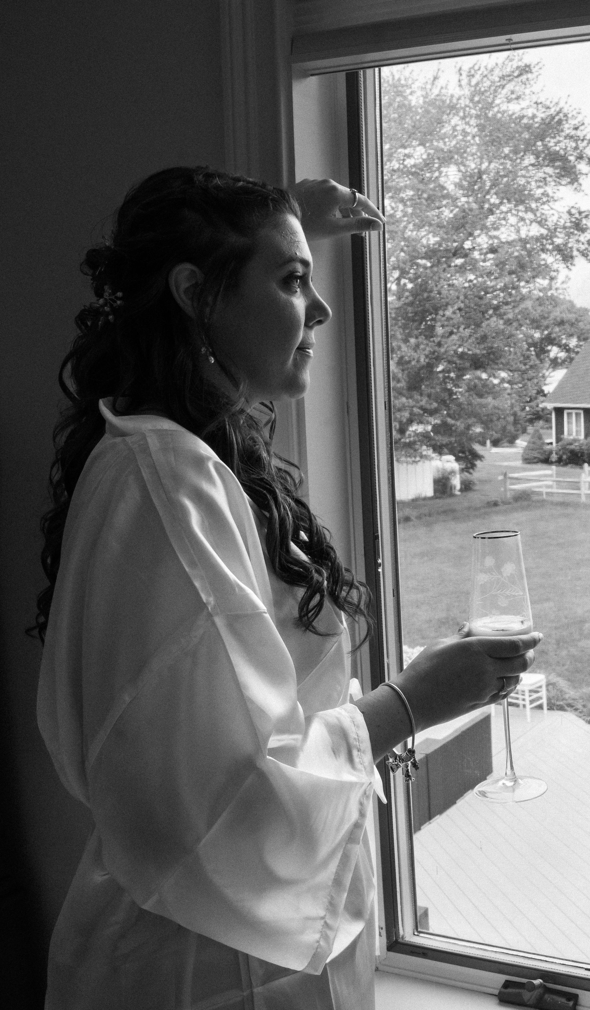 A person in a white robe holds a glass of wine while standing by a window and looking outside.
