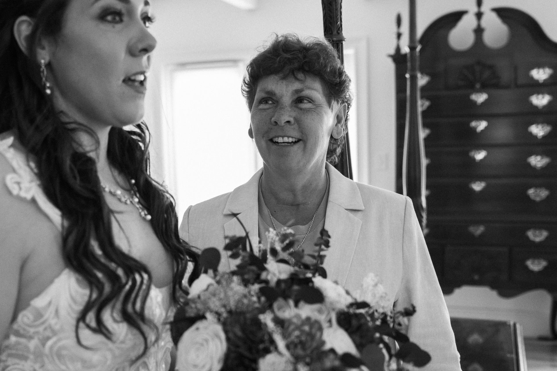 A woman in a bridal dress looks toward an older woman, who smiles at her, in a room with a tall wooden cabinet.