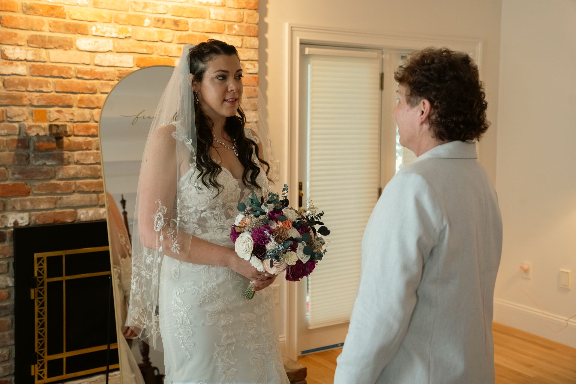 A bride in a white lace dress and veil holds a bouquet while looking at a person in a light blue suit.