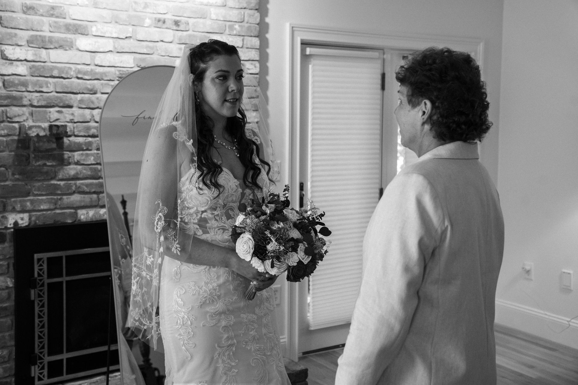 A bride in a lace wedding dress and veil stands indoors holding a bouquet while talking to a person in a blazer.