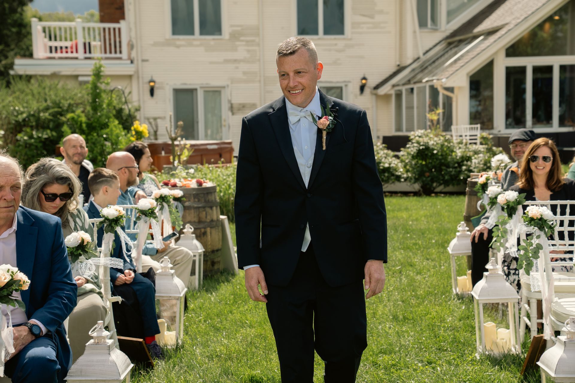 A person in a suit walks down a grassy aisle at an outdoor wedding, passing rows of guests seated in white chairs.
