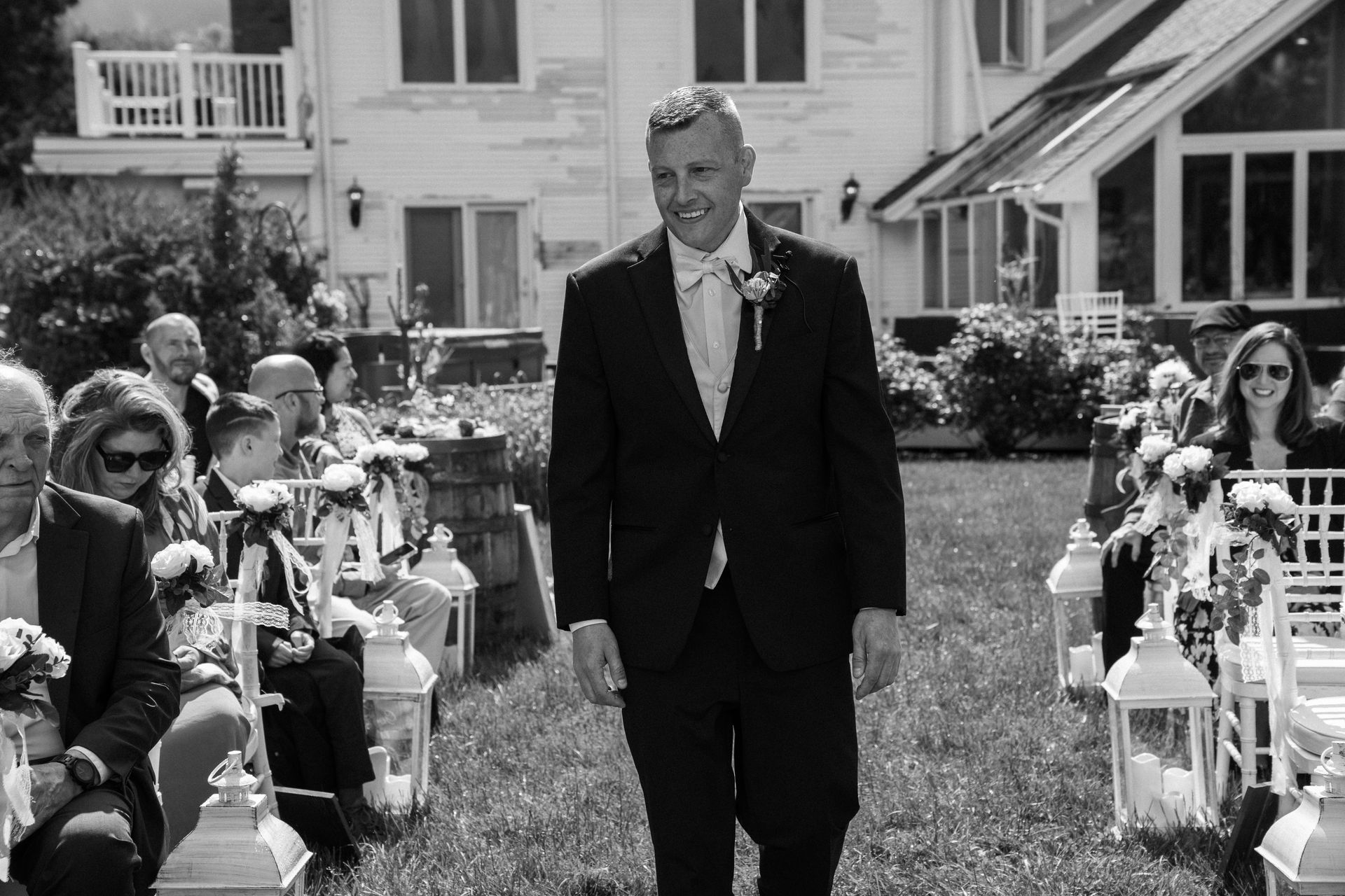 A person in a suit walks down an outdoor wedding aisle, smiling, past rows of seated guests and white lanterns.