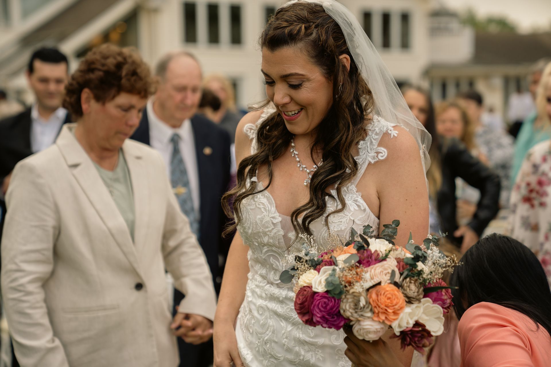 A bride in a white wedding gown holding a colorful bouquet, surrounded by guests at an outdoor event.