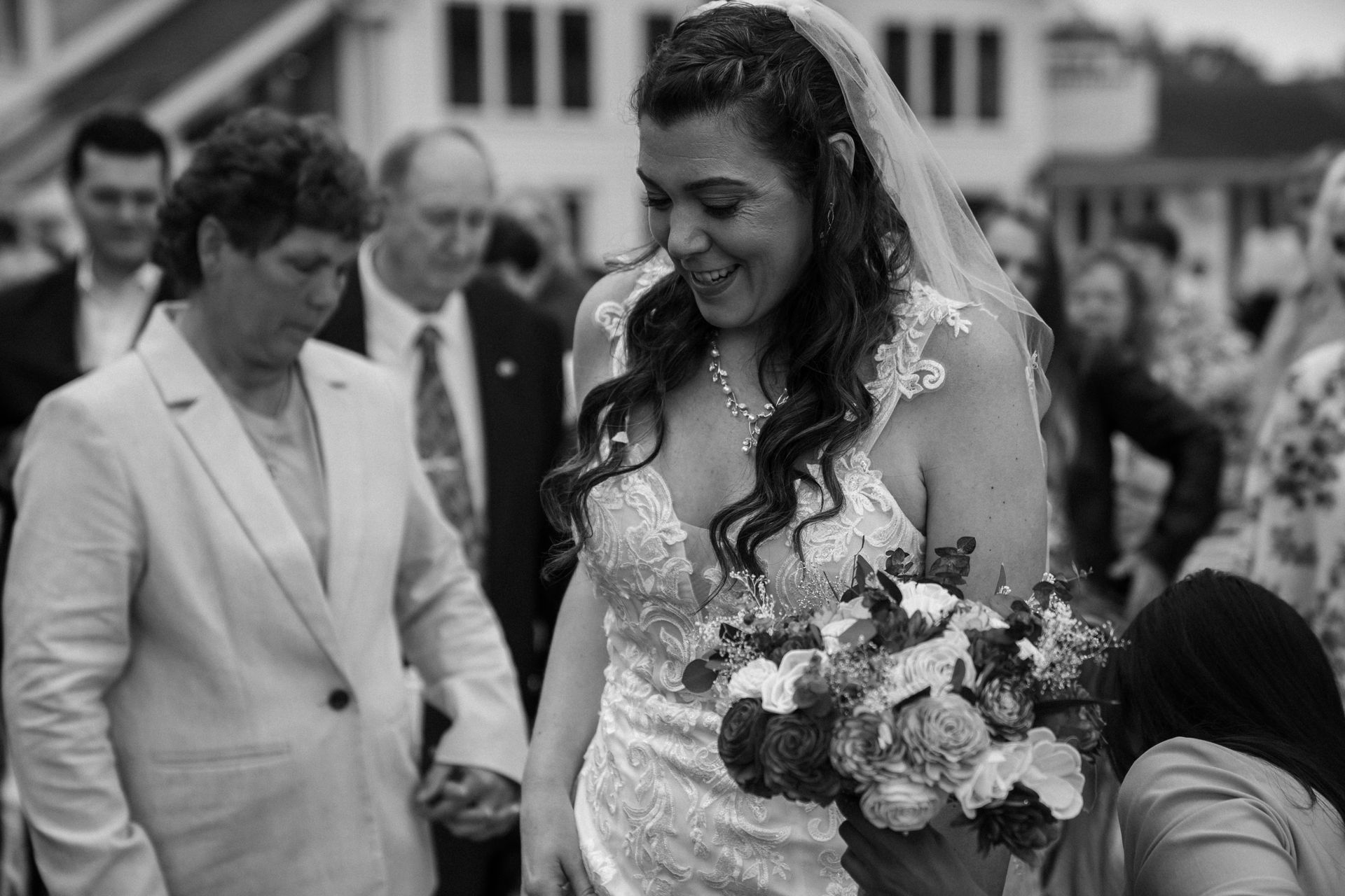 A bride in a white wedding gown holding a bouquet smiles, walking outdoors with family members nearby.