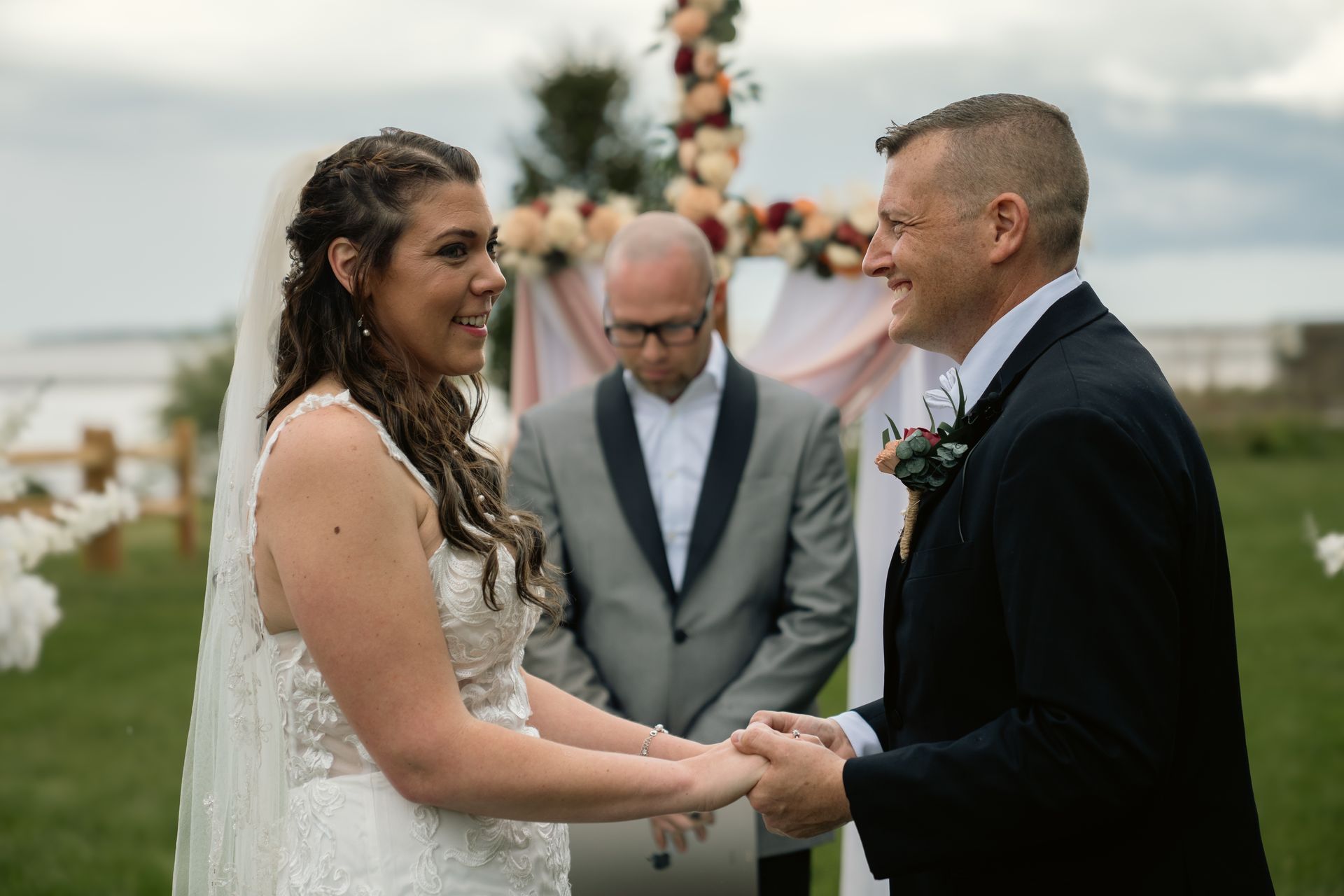 A bride and groom holding hands and smiling at each other during an outdoor wedding ceremony with an officiant behind them.