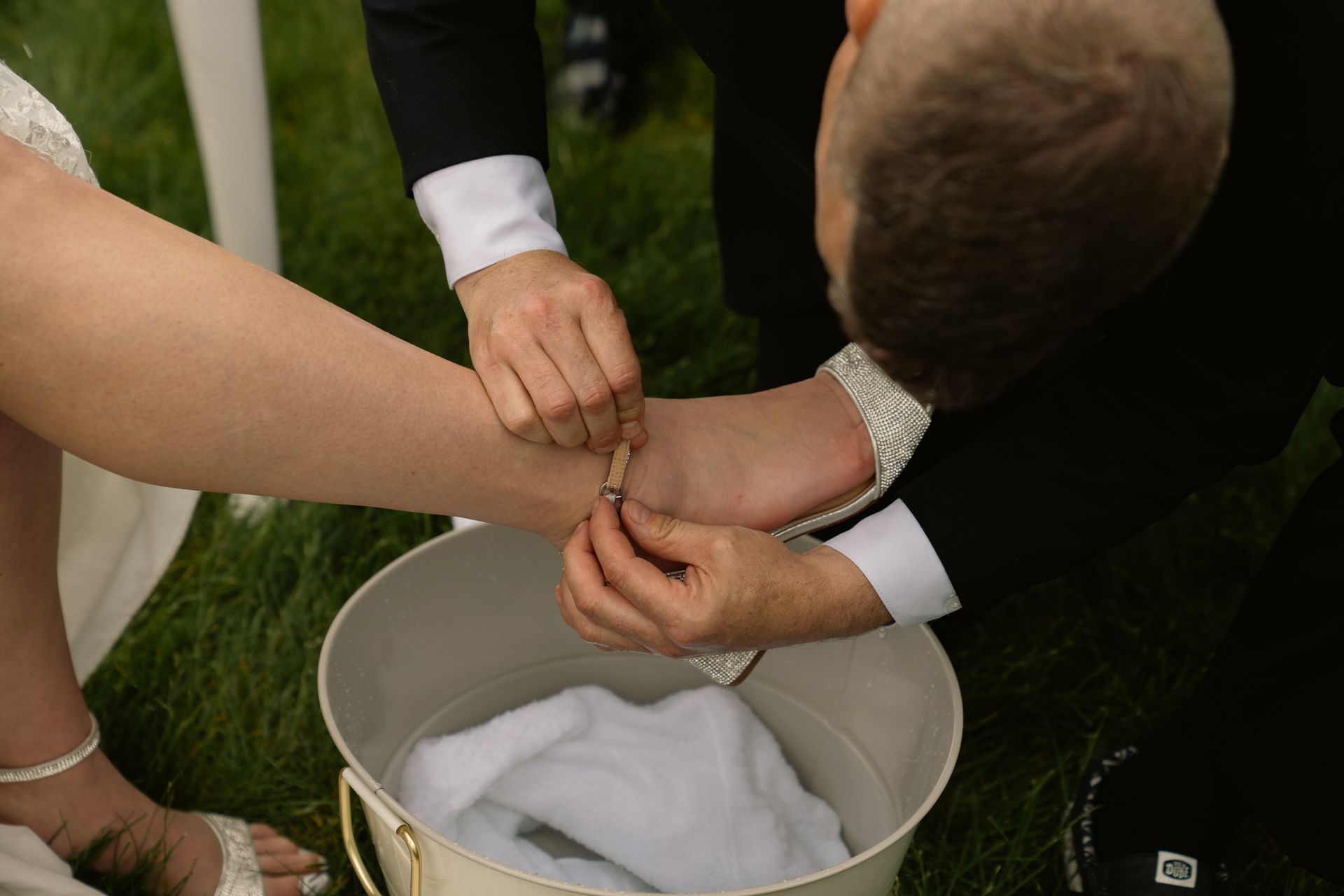 A person in a suit kneels to help fasten the ankle strap of a bride's embellished heel over a water basin on the grass.